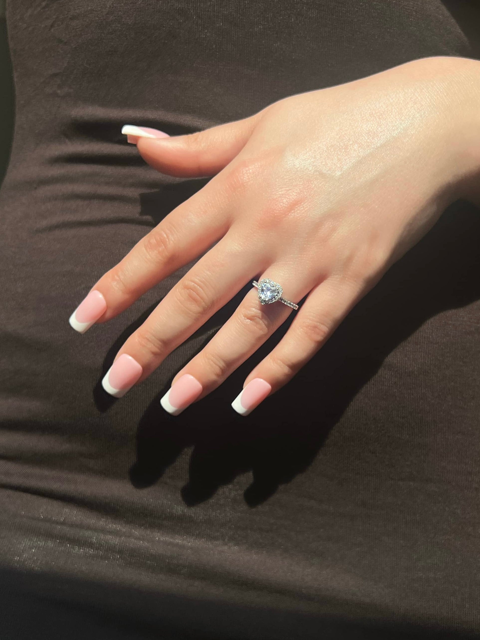 A woman’s hand with white-tip square-shaped acrylic nails displays a stunning silver heart-shaped diamond at the center, and paved stones on the sides. The ring is elegantly placed on her finger, perfect for expressing love. She is wearing a brown dress
