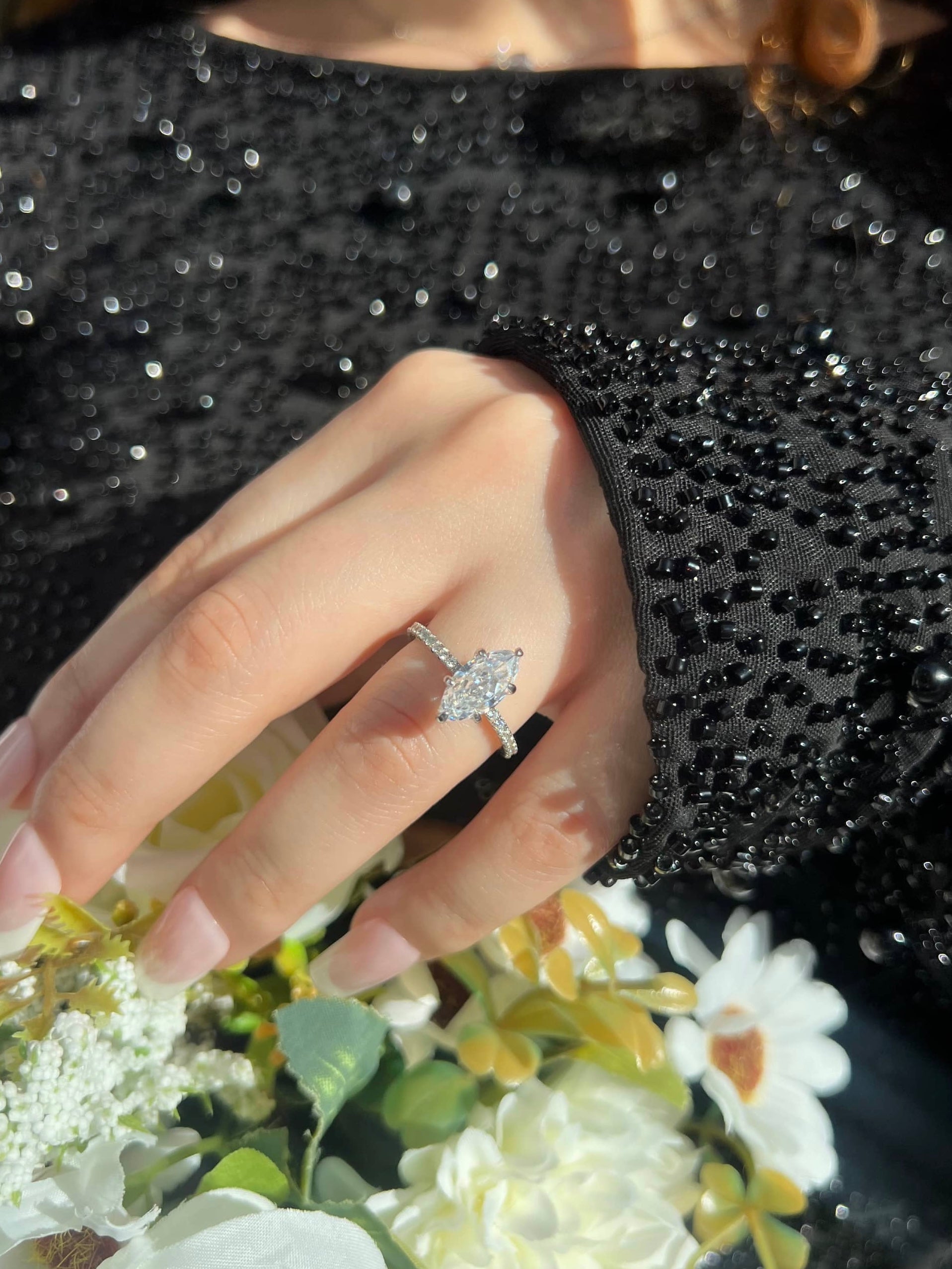 A close-up of a woman's hand wearing a silver engagement ring featuring a large marquise-shaped diamond at the center, surrounded by smaller diamonds on the band. Her hand rests over a bouquet of white flowers. She is wearing a black embellished dress