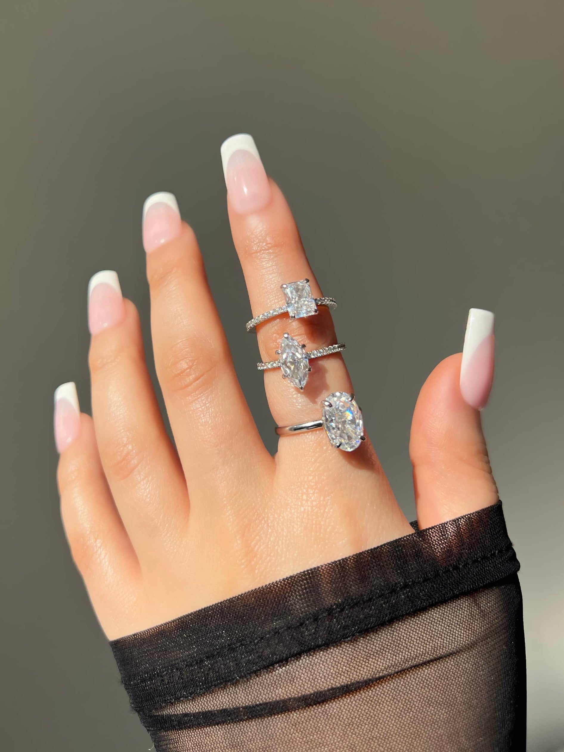 A close-up of a woman's hands showcasing French manicure nails. Three silver rings adorn her index finger. The first ring is a radiant cut, the second is a marquise cut and the third is an oval cut solitaire.