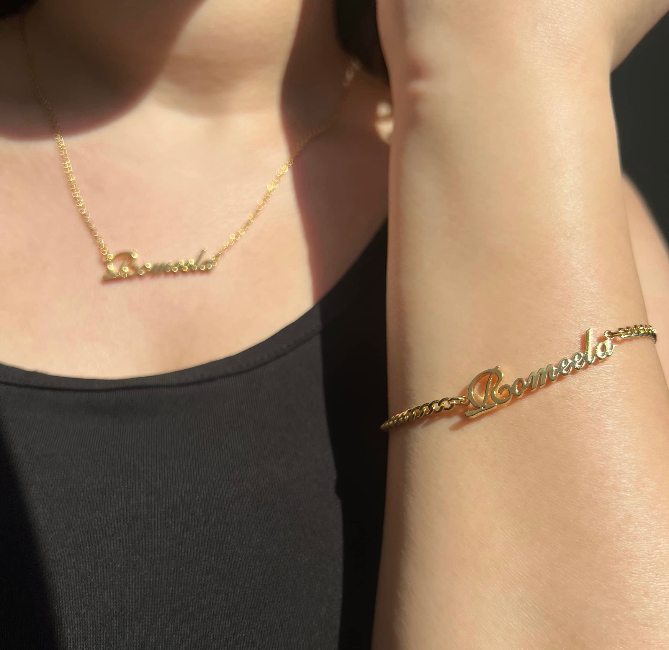 A woman wearing a black top, showcasing a gold necklace and bracelet. Both pieces feature the name 'Romeela' beautifully inscribed, adding a personal touch to her look. The jewellery shines elegantly, highlighting her unique style and individuality