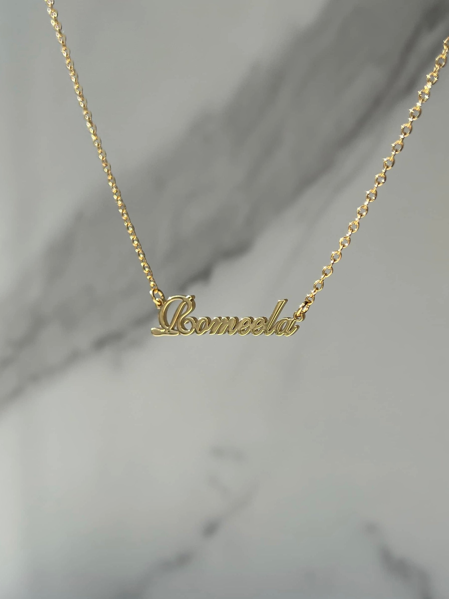 A gold necklace featuring a delicate nameplate pendant that reads 'Romeela' in fancy cursive script. The necklace is elegantly displayed on a sleek marble background, highlighting its shine and beauty, making it a perfect piece for personal expression