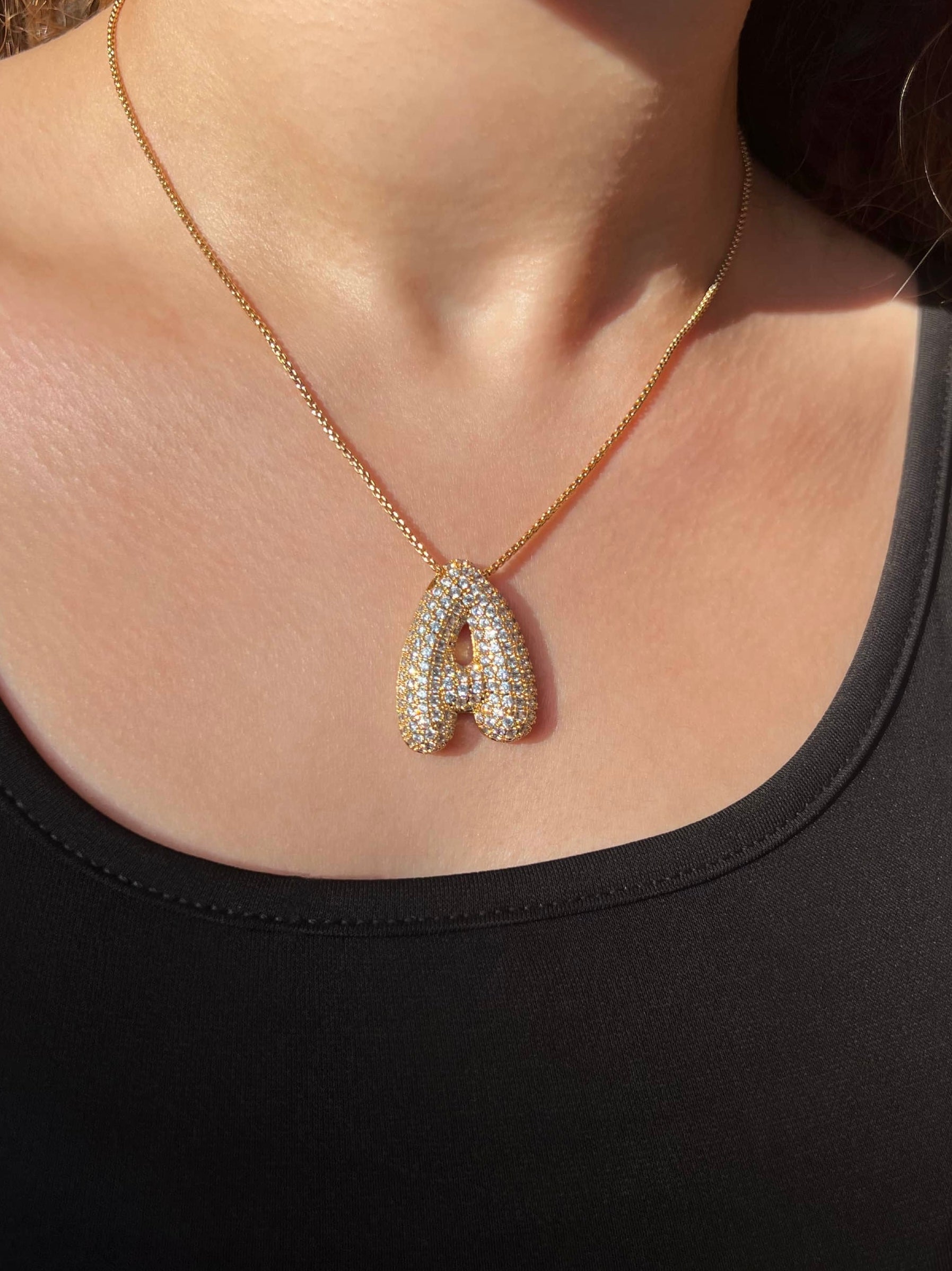 A woman wearing a stylish black top paired with a gold chain necklace. The necklace features a pendant shaped like the letter 'A', beautifully encrusted with small, clear stones that add a touch of sparkle. The combination creates a chic and elegant look