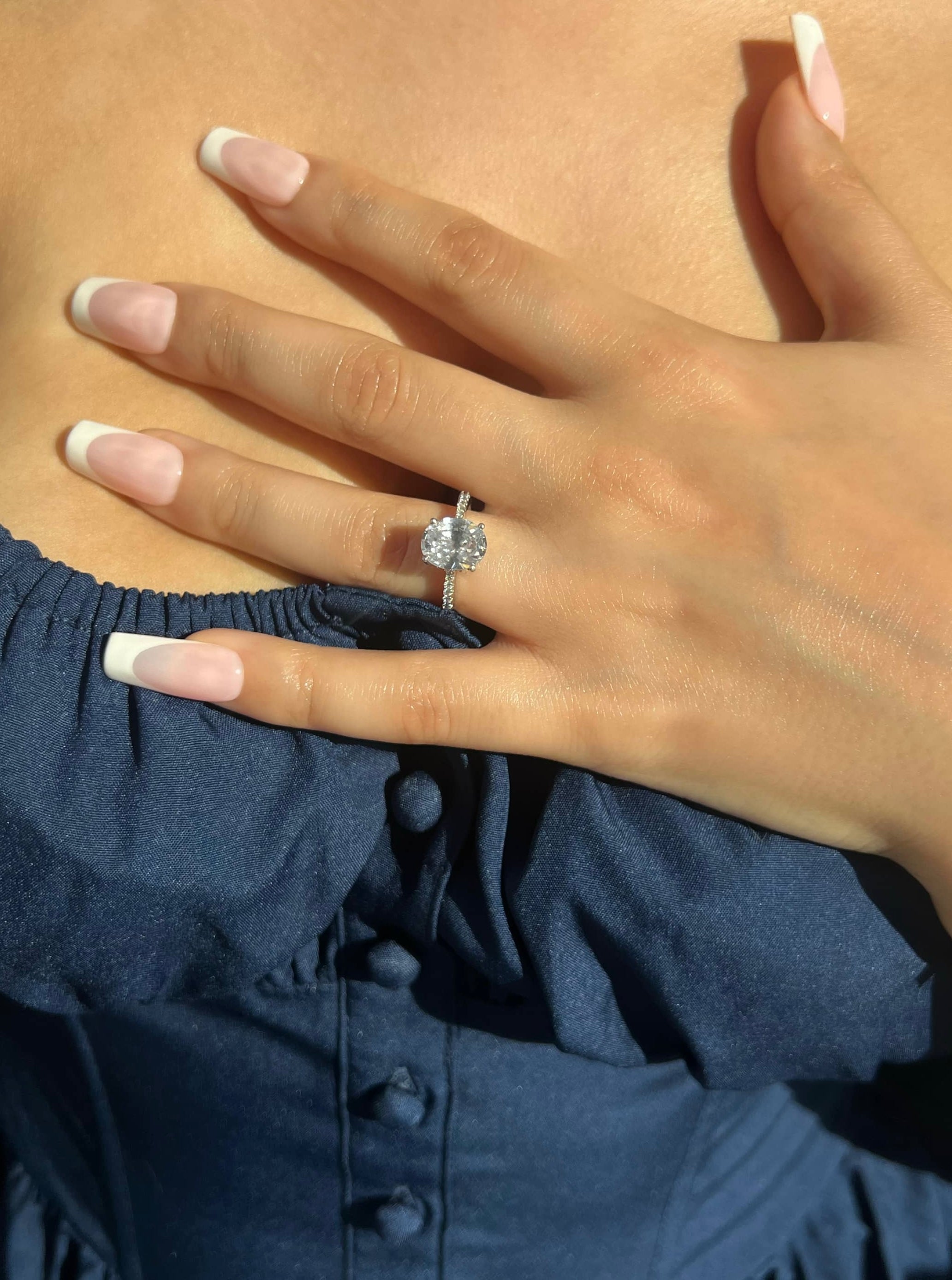 A woman’s hand with French manicured nails showcases a stunning silver ring featuring a 2 carat 8*10mm oval-shaped diamond surrounded by a line of smaller stones. The hand is gently resting on a navy blue dress, highlighting the ring's beauty and sparkle