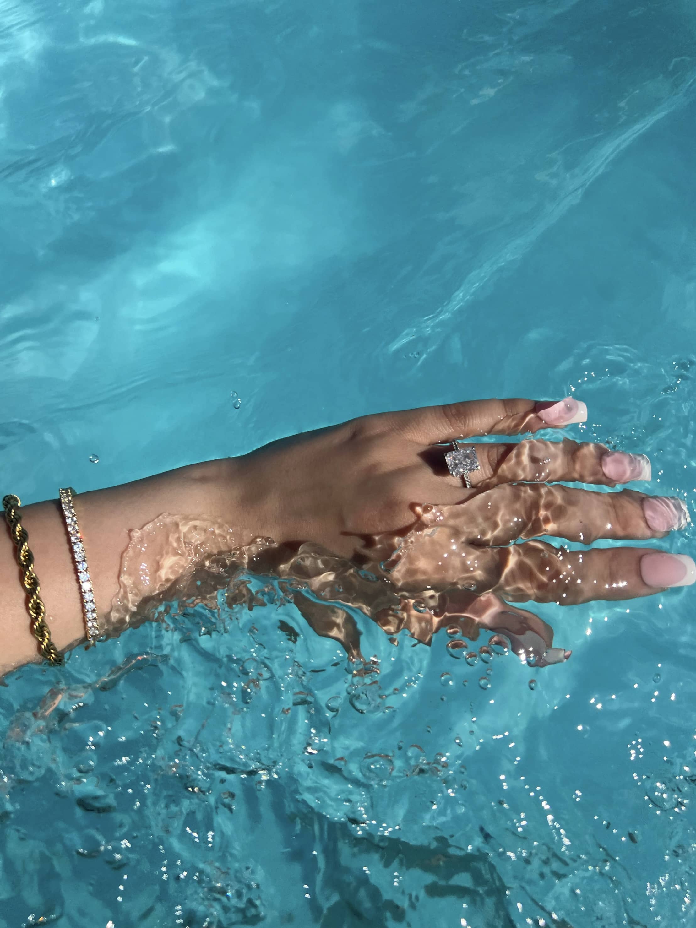 A woman’s hand with long, French tip nails showcases a radiant cut diamond ring surrounded by smaller diamonds, submerged in clear blue water. A gold rope chain bracelet with diamonds adorns the wrist, creating an elegant and luxurious look.