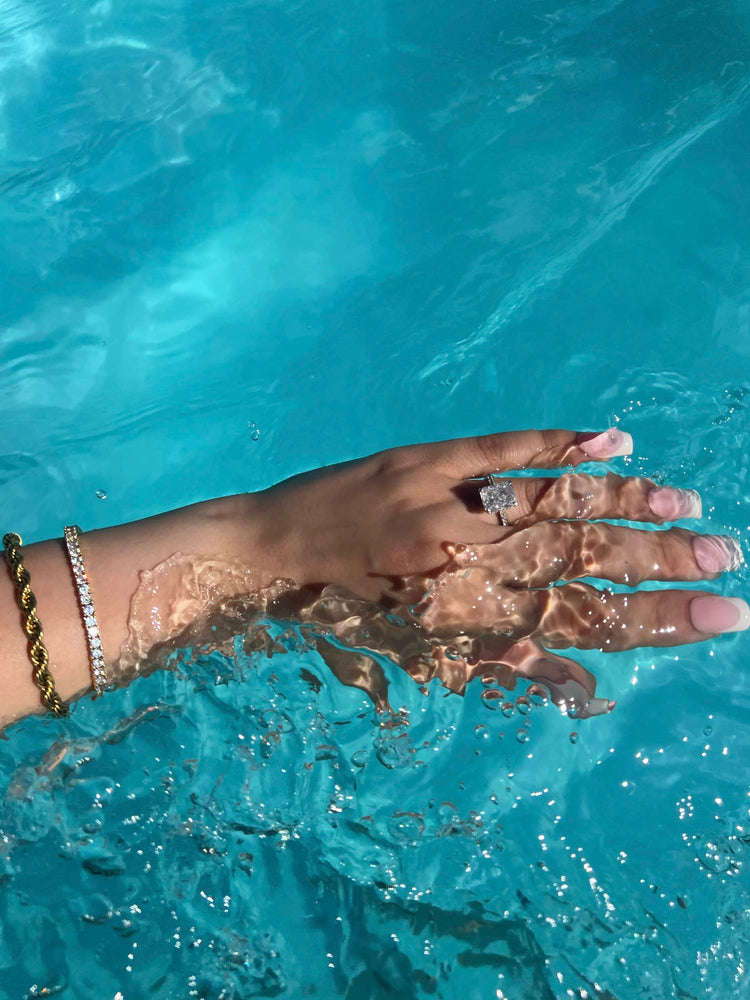 A woman’s hand with long, French tip nails showcases a radiant cut diamond ring surrounded by smaller diamonds, submerged in clear blue water. A gold rope chain bracelet with diamonds adorns the wrist, creating an elegant and luxurious look.