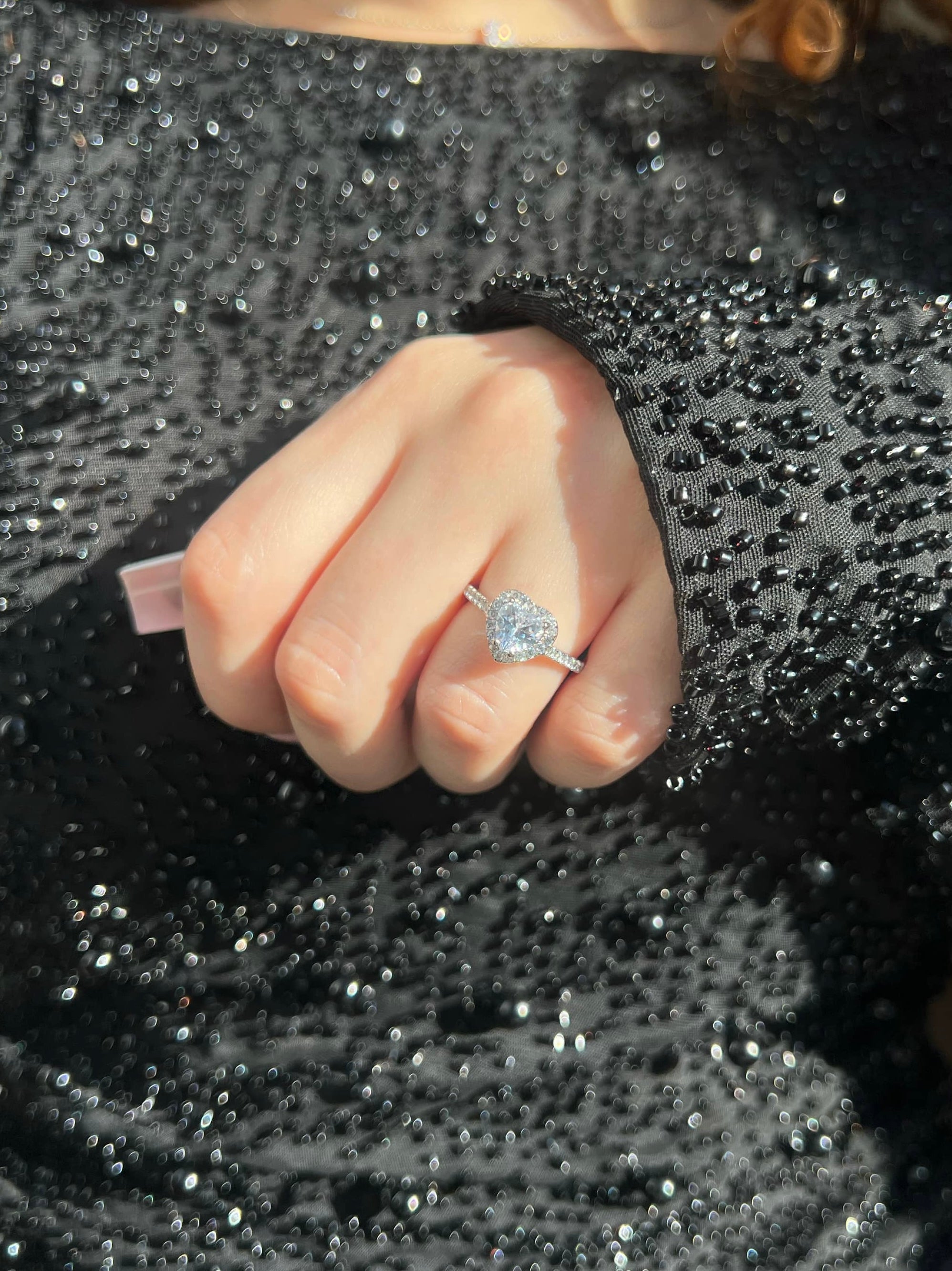 A woman's hand adorned with a stunning silver ring on her ring finger, featuring a large heart-shaped diamond in the center, surrounded by a halo of smaller diamonds, complementing her black pearl embellished dress, adding a touch of sparkle.