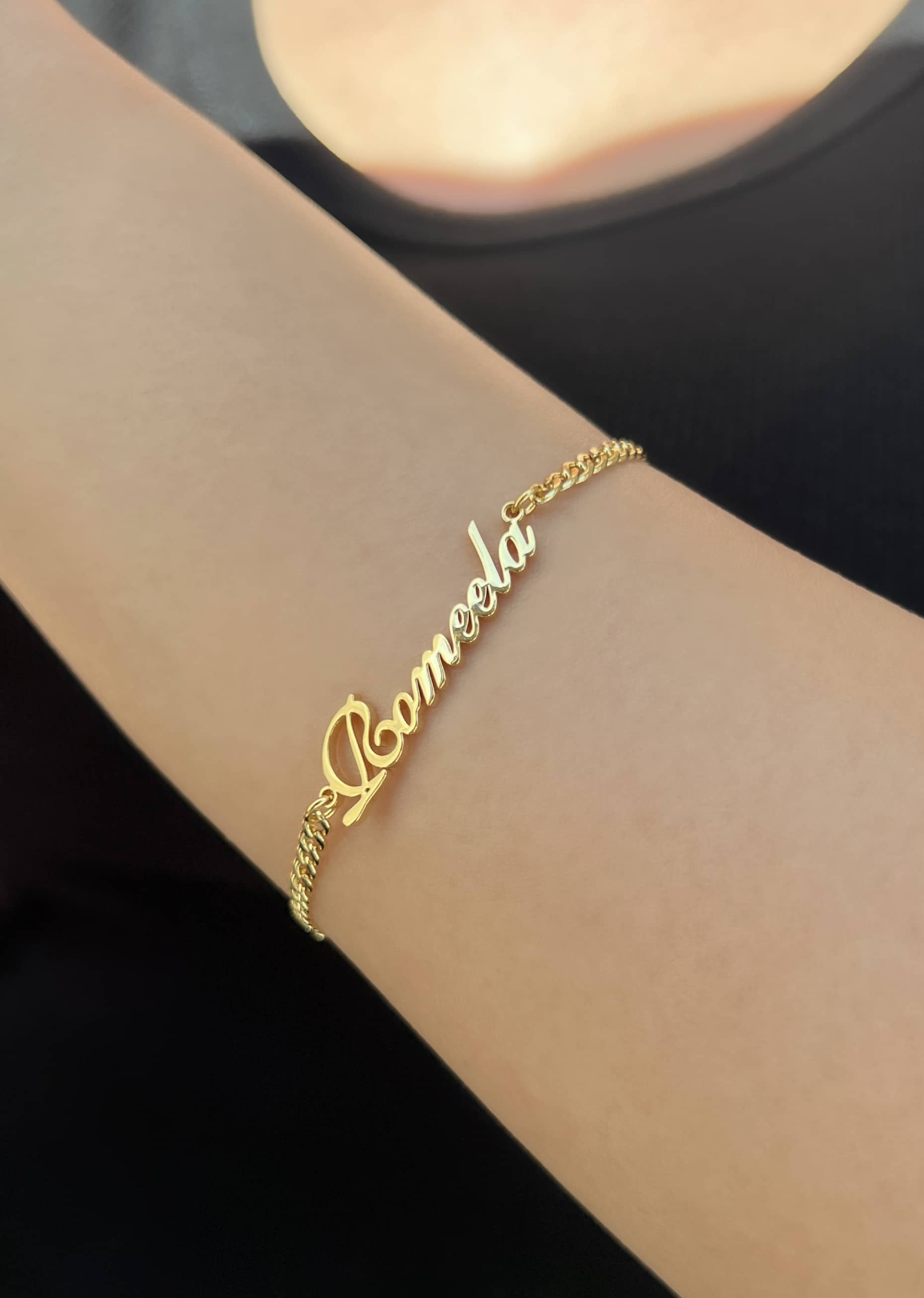 A gold bracelet featuring the name 'Romeela'. The bracelet is styled with a classic curb chain, adding a touch of sophistication. It's worn on a woman’s wrist, showcasing its beauty and personal significance as a cherished accessory
