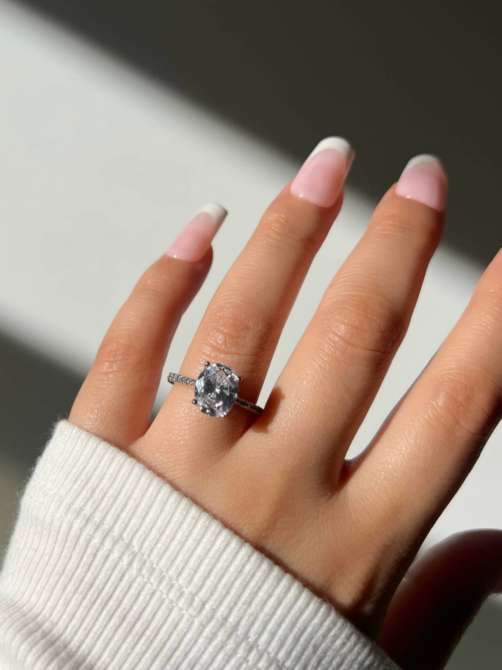 A woman’s hand with white French tip nails featuring a large 2CT 8*10mm silver oval-shaped diamond at the center, surrounded by smaller diamonds in a halo setting. The ring is elegantly placed on her ring finger, highlighting its brilliance and sparkle
