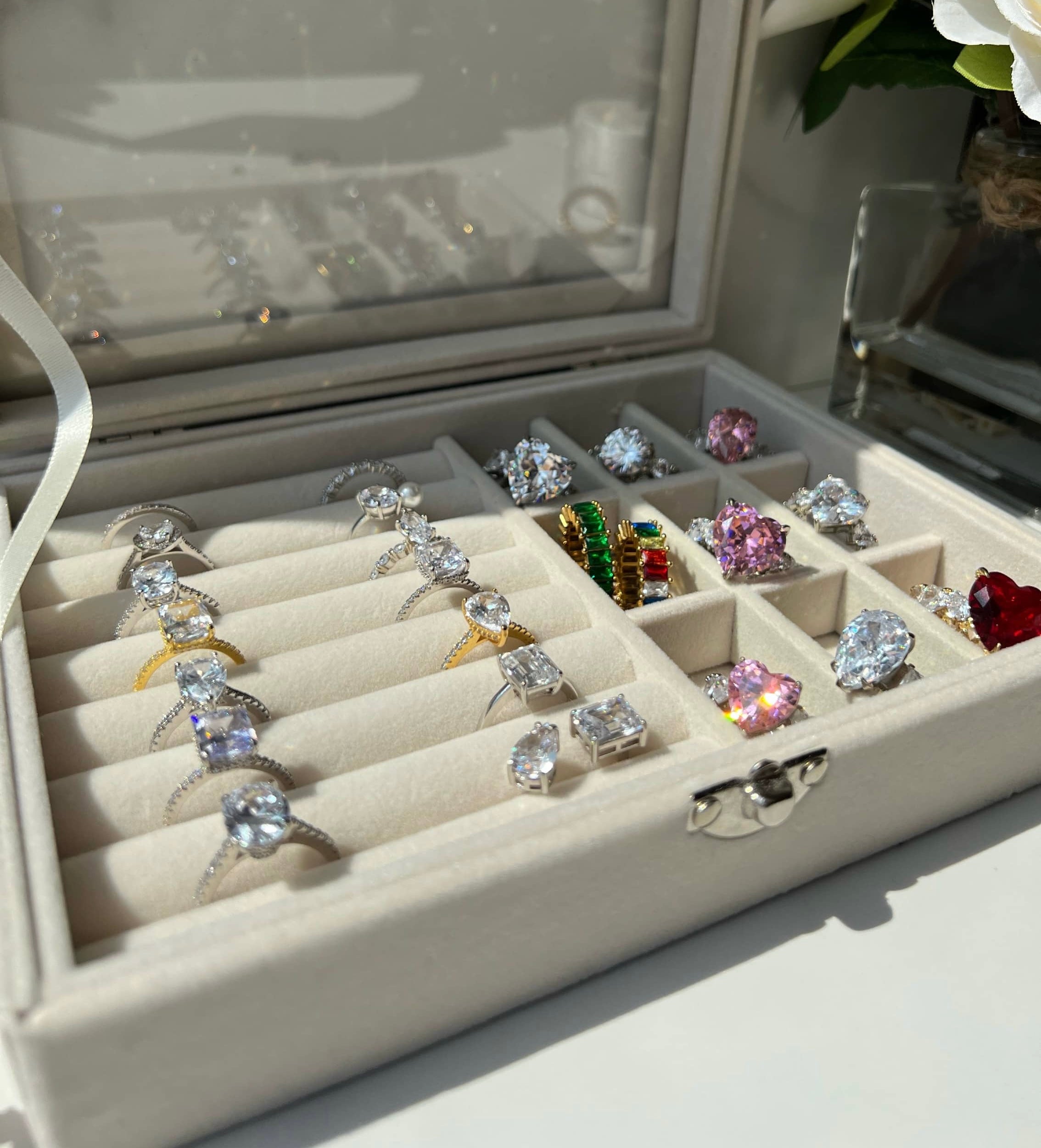 A white velvet jewelry box showcasing an array of stunning rings, including gold diamond rings, a a colourful rainbow ring, pink heart-shaped diamond rings, and diamond simulant engagement rings, all beautifully arranged in a neat grid pattern.