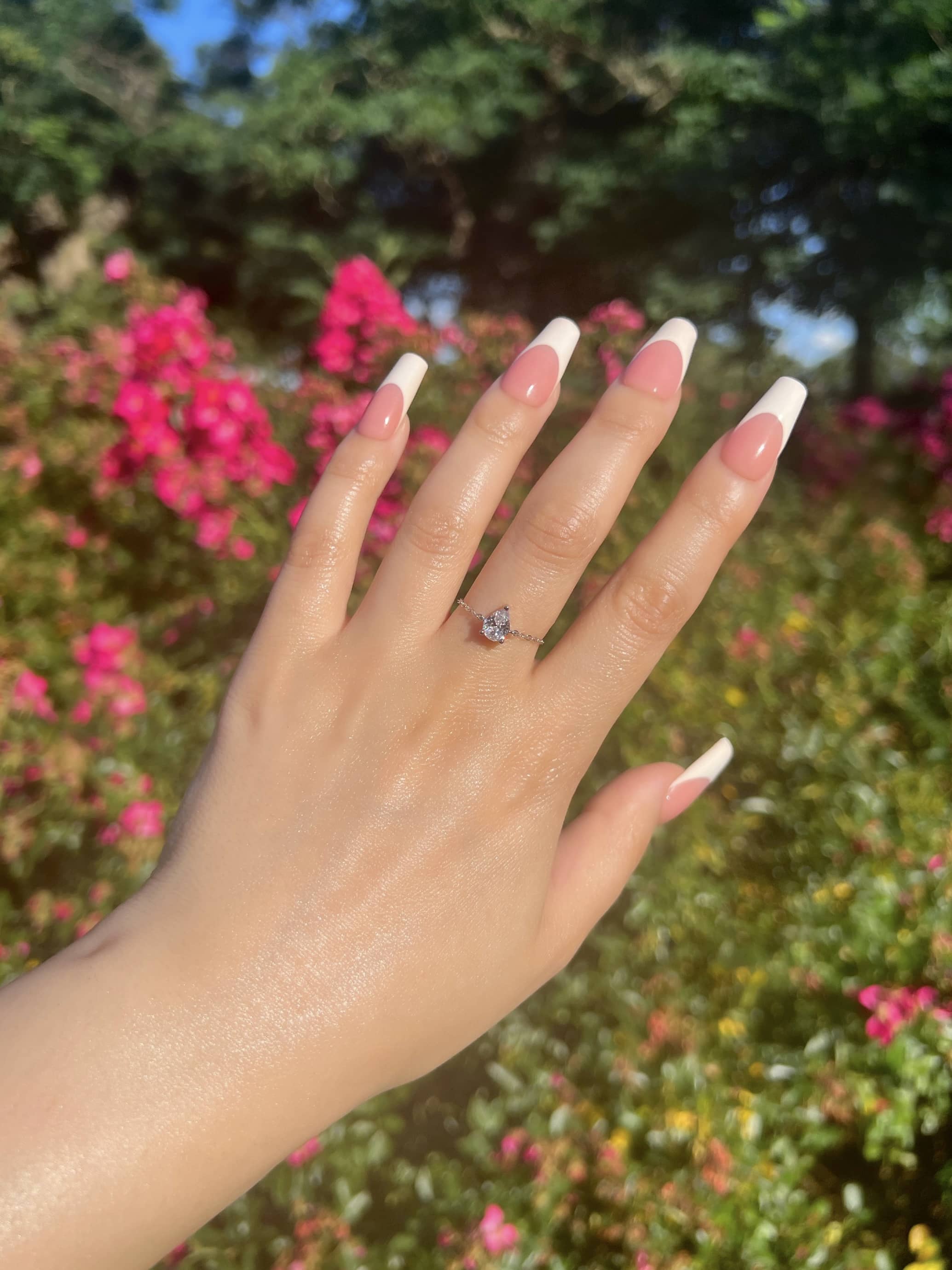 A woman’s hand with French manicured nails showcases a stunning pear-shaped diamond chain ring. The hand is elegantly posed against a blurred background of pink flowers and lush green foliage, adding a soft, romantic touch to the overall aesthetic.
