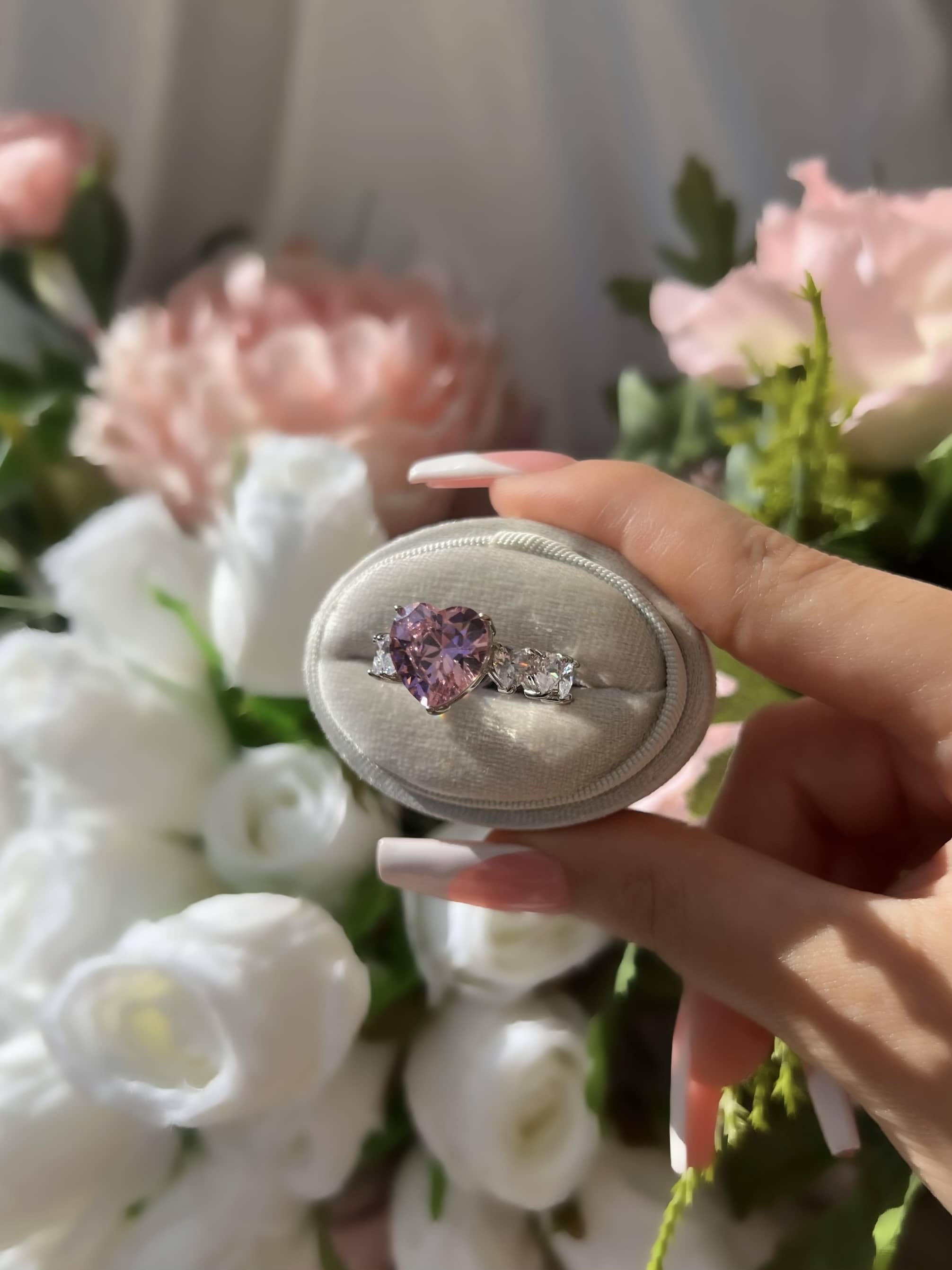 A woman’s hand holding an oval-shaped ring box containing a silver ring with a large heart-shaped pink stone at the center, surrounded by smaller clear stones. The ring is beautifully complemented by white & pink flowers with green stems in the background