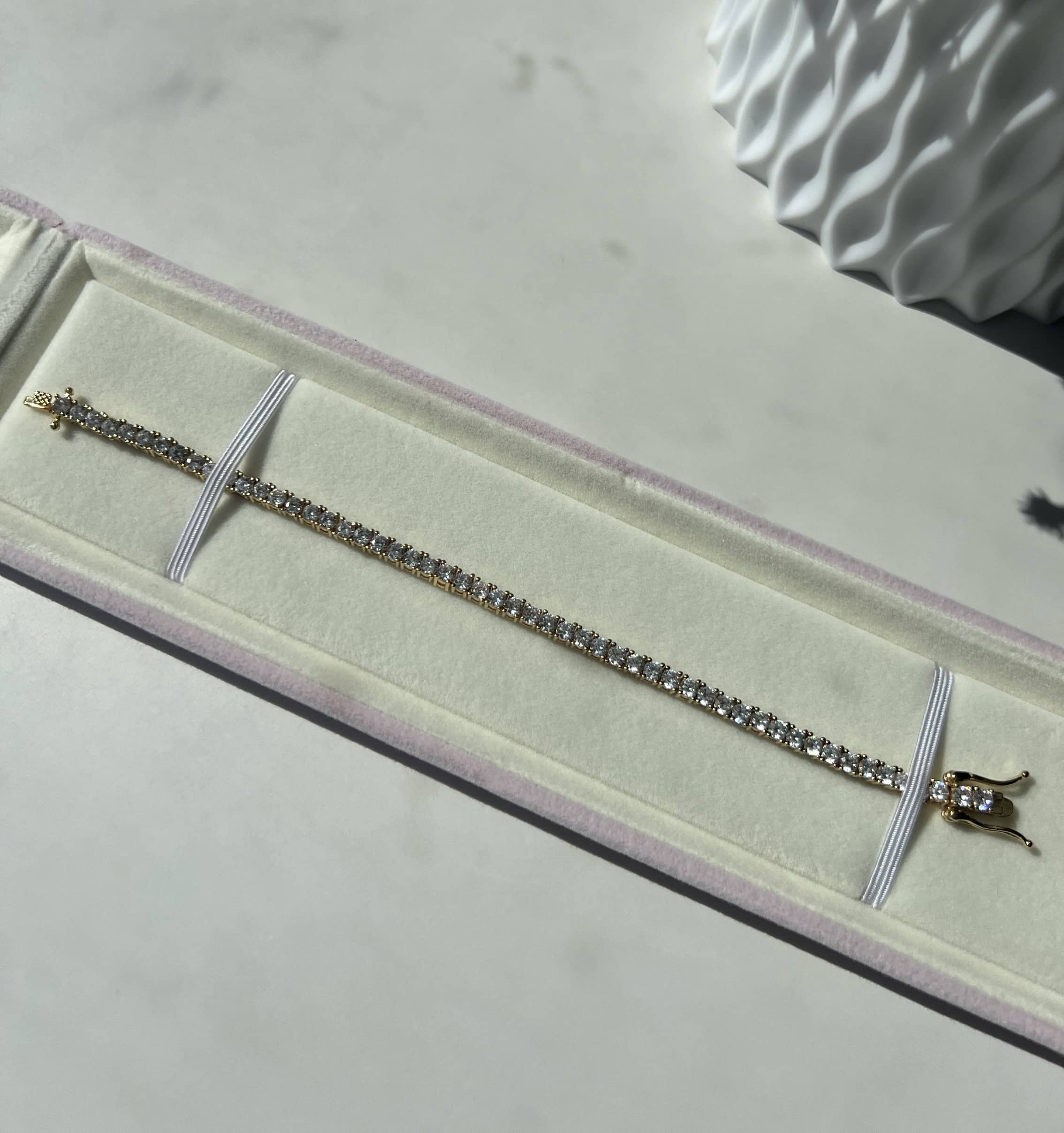 Elegant gold and diamond tennis bracelet displayed in a white velvet box with pink trim. The bracelet features small, clear diamonds set in a gold setting, showcasing its exquisite craftsmanship and sparkle, with the clasp facing right.