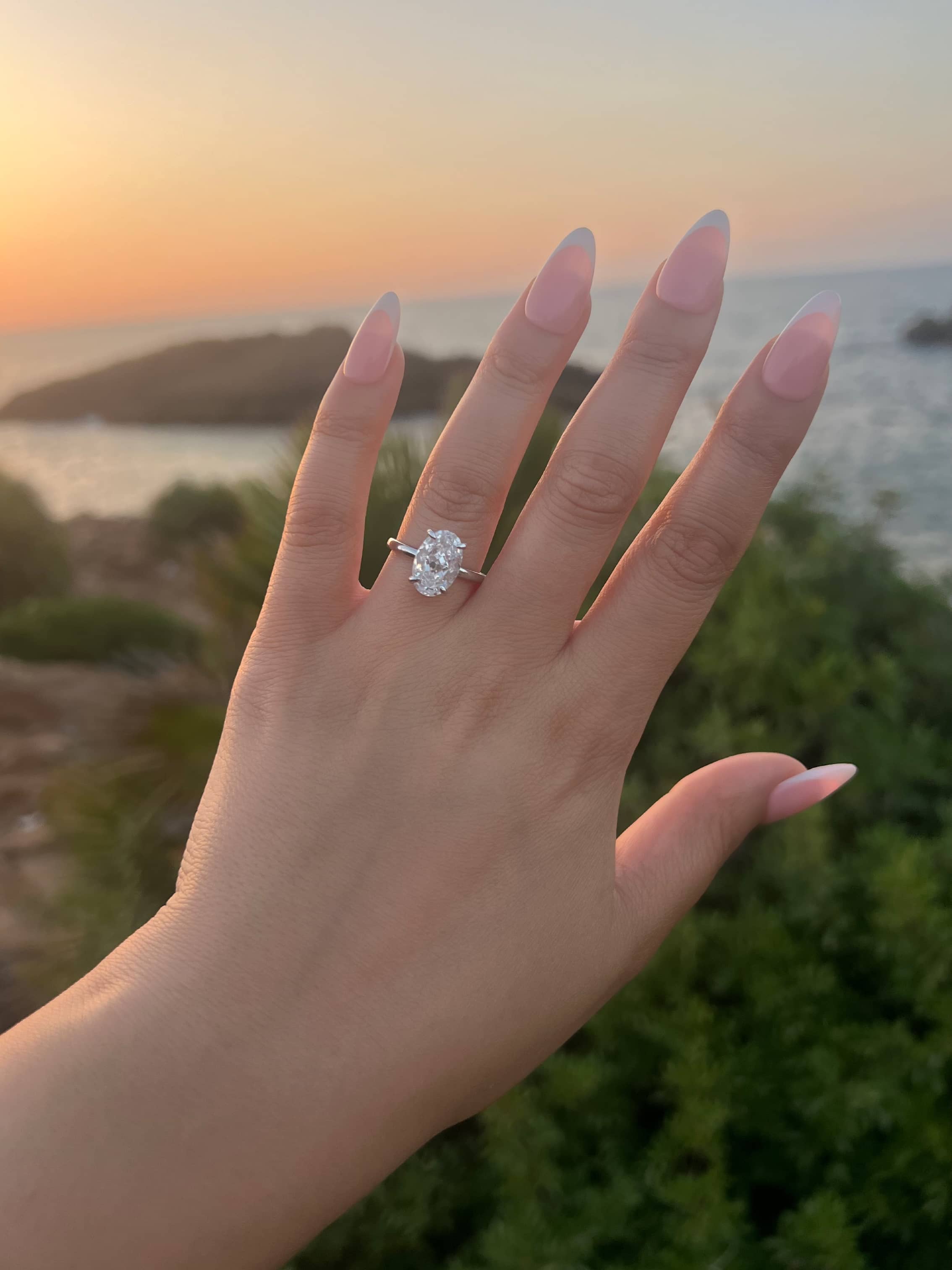 A woman’s hand with almond-shaped nails showcases a silver band with an oval-shaped diamond ring, set against a blurred sunset backdrop over water and greenery, highlighting the elegant solitaire setting of the ring