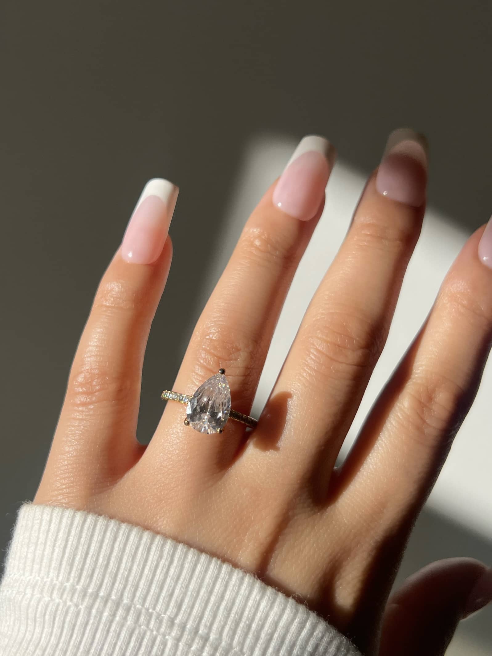 A woman’s hand adorned with a stunning pear-shaped diamond engagement ring with pave stones on both sides, on her ring finger. Her hand features a long, square french manicure and a white rib sweater, showcasing an elegant and luxury style