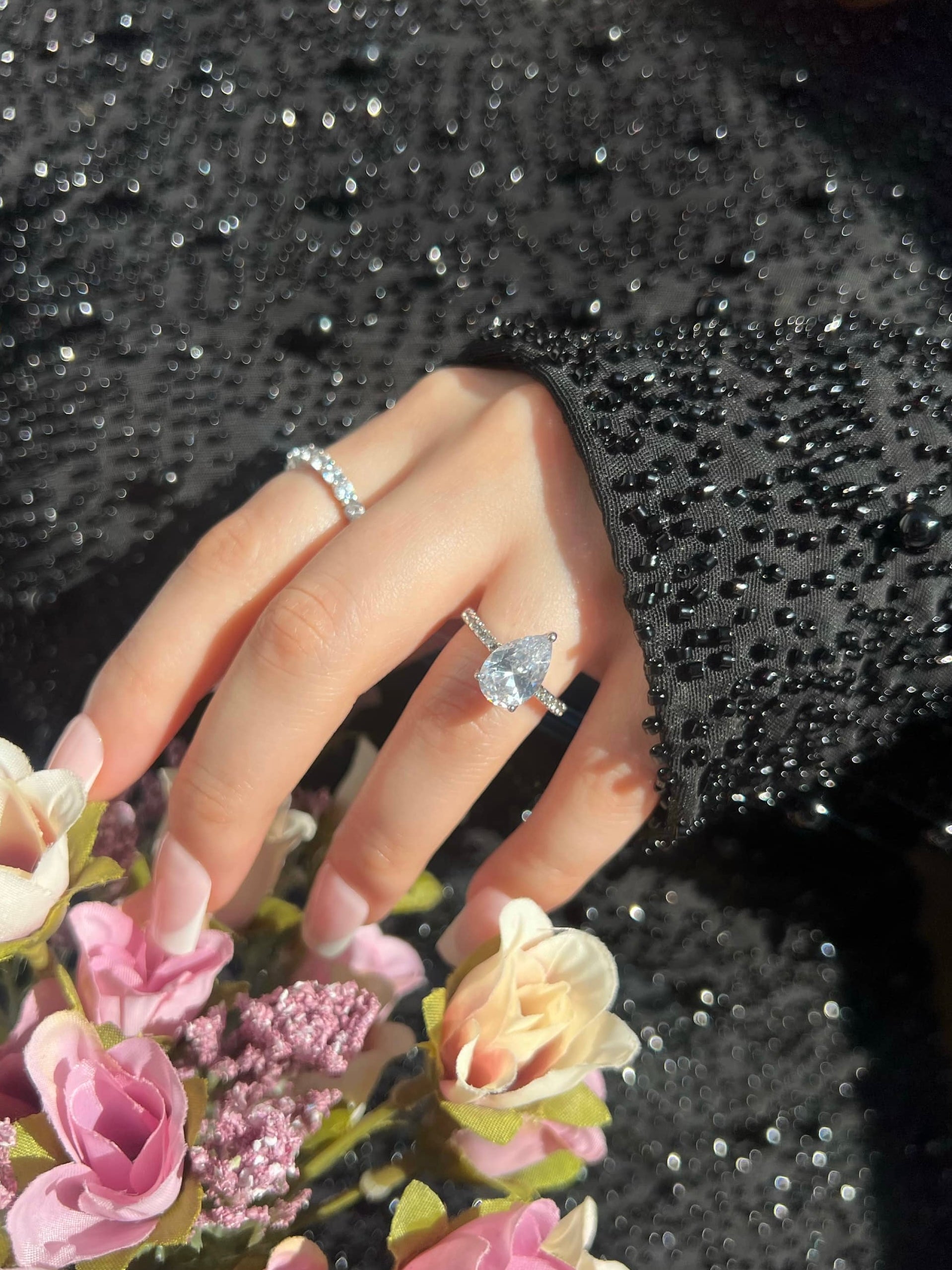 A woman’s hand showcasing a pear-shaped engagement ring on her ring finger, surrounded by a thin band of smaller diamonds and a smaller band ring on her index finger, with several pink roses in the background. She is wearing a black embellished dress