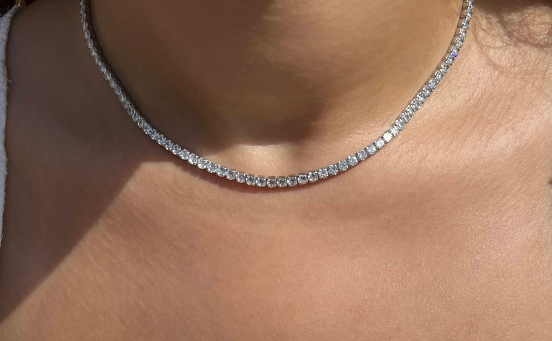An image of a woman wearing a sparkling tennis necklace with clear stones, elegantly draped over a white top. The necklace showcases a modern and luxurious design, adding a touch of timeless sophistication to the overall look. Perfect for any occasion