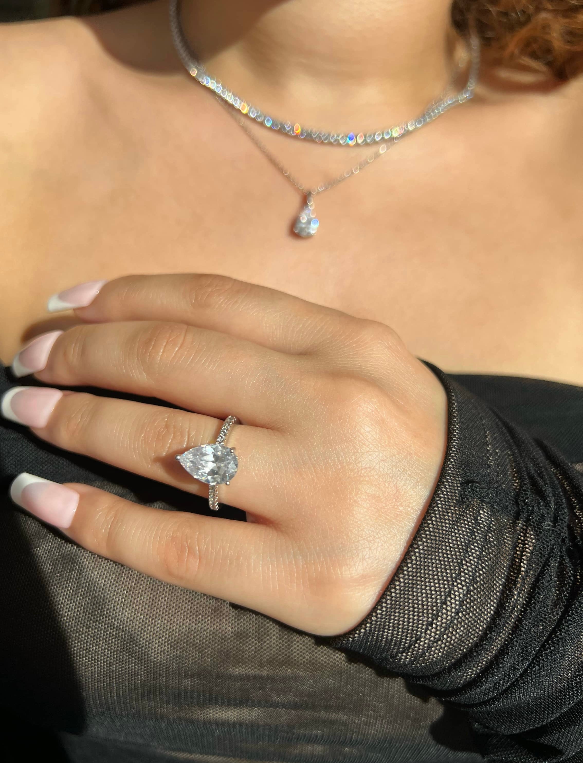 A close-up of a woman’s hand adorned with a stunning silver pear-shaped diamond engagement ring featuring a pave band of smaller diamonds. Her hand showcases a french manicure, complemented by a diamond pear cut pendant necklace and tennis necklace