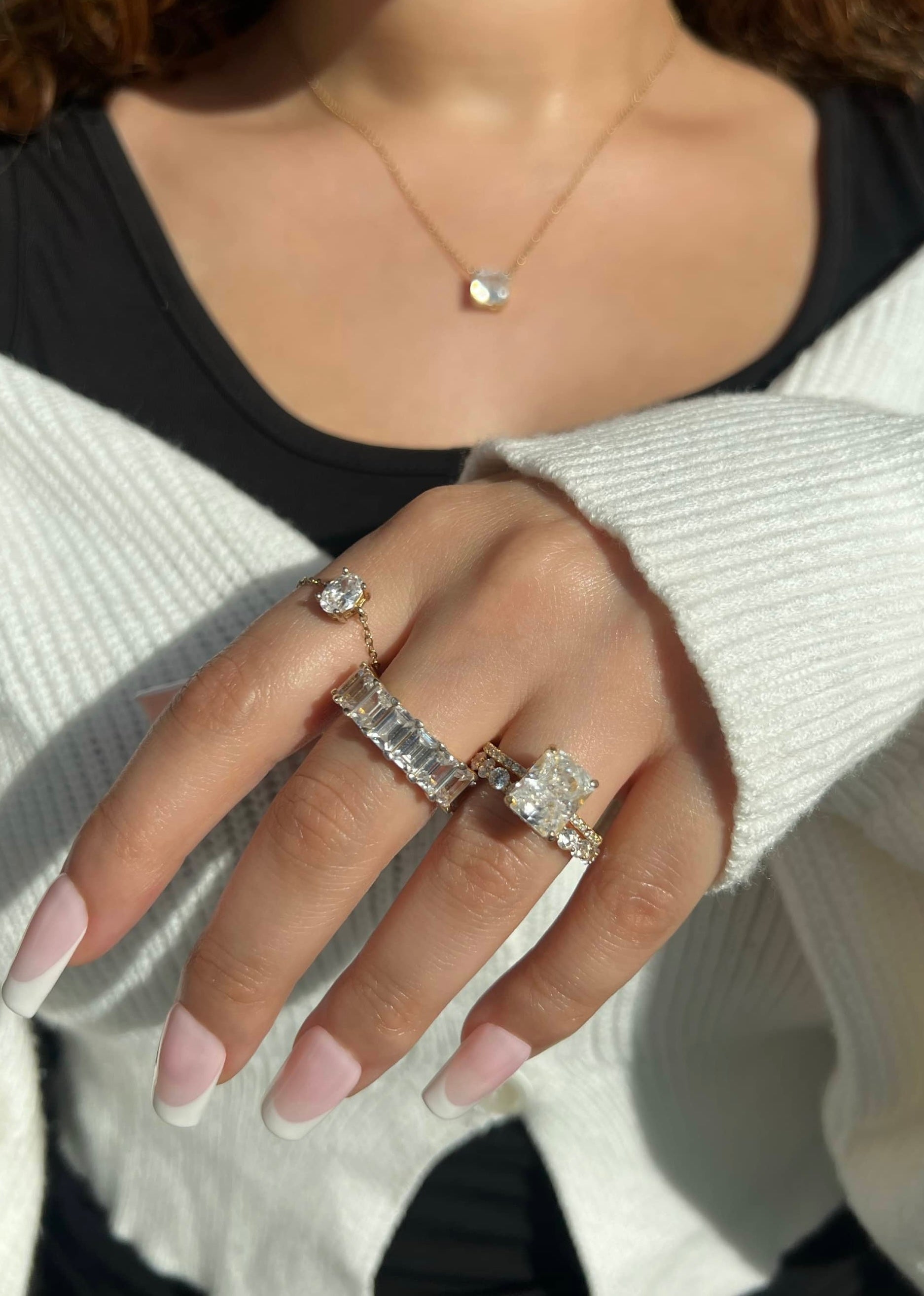 A woman’s hand adorned with four stunning gold diamond rings: a thick emerald cut band, a round cut band, a large radiant-cut diamond, and a small chain ring. The hand also features a gold chain with a diamond pendant,
complemented by a French manicure.