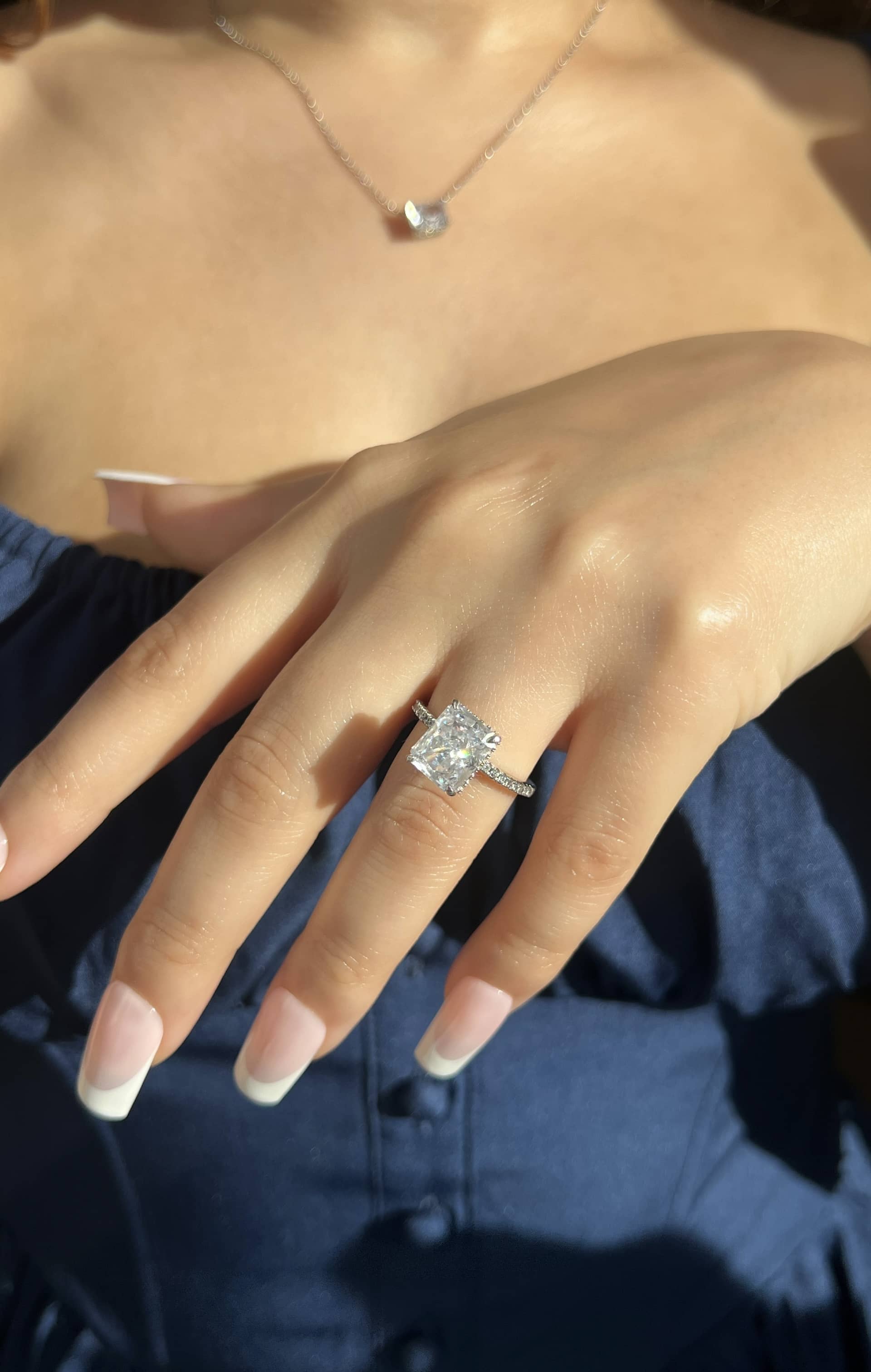 A woman’s hand adorned with a stunning silver radiant-shaped diamond ring on her ring finger, complemented by a delicate silver chain necklace around her neck and a navy blue corset top. She has French manicured nails, exuding elegance and sophistication