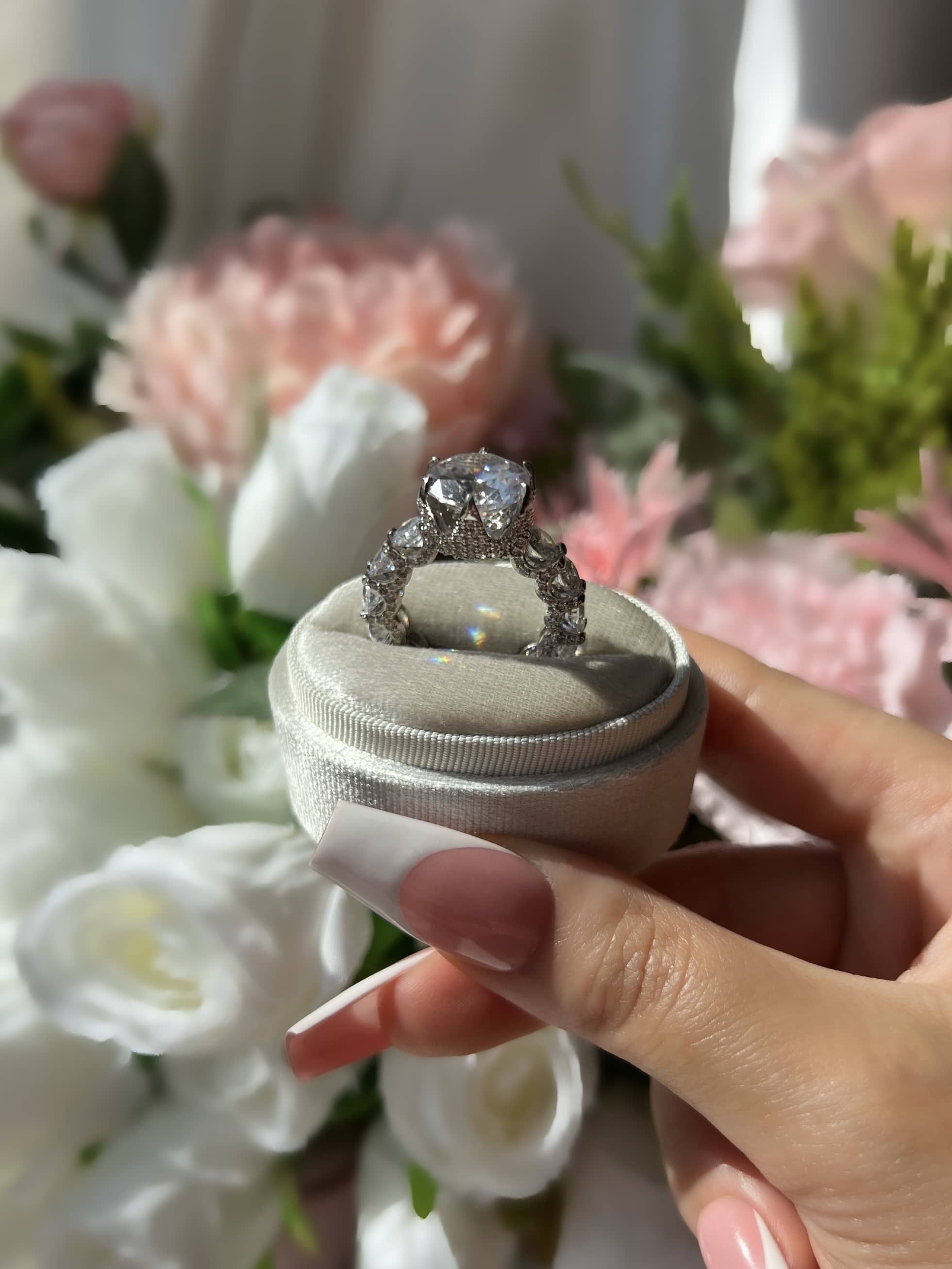 A woman’s hand with long french tip nails holds a ring box containing a stunning round diamond ring, surrounded by smaller diamonds. The hand rests against a bouquet of pink and white flowers, emphasizing the elegance of the ring in a beautiful setting