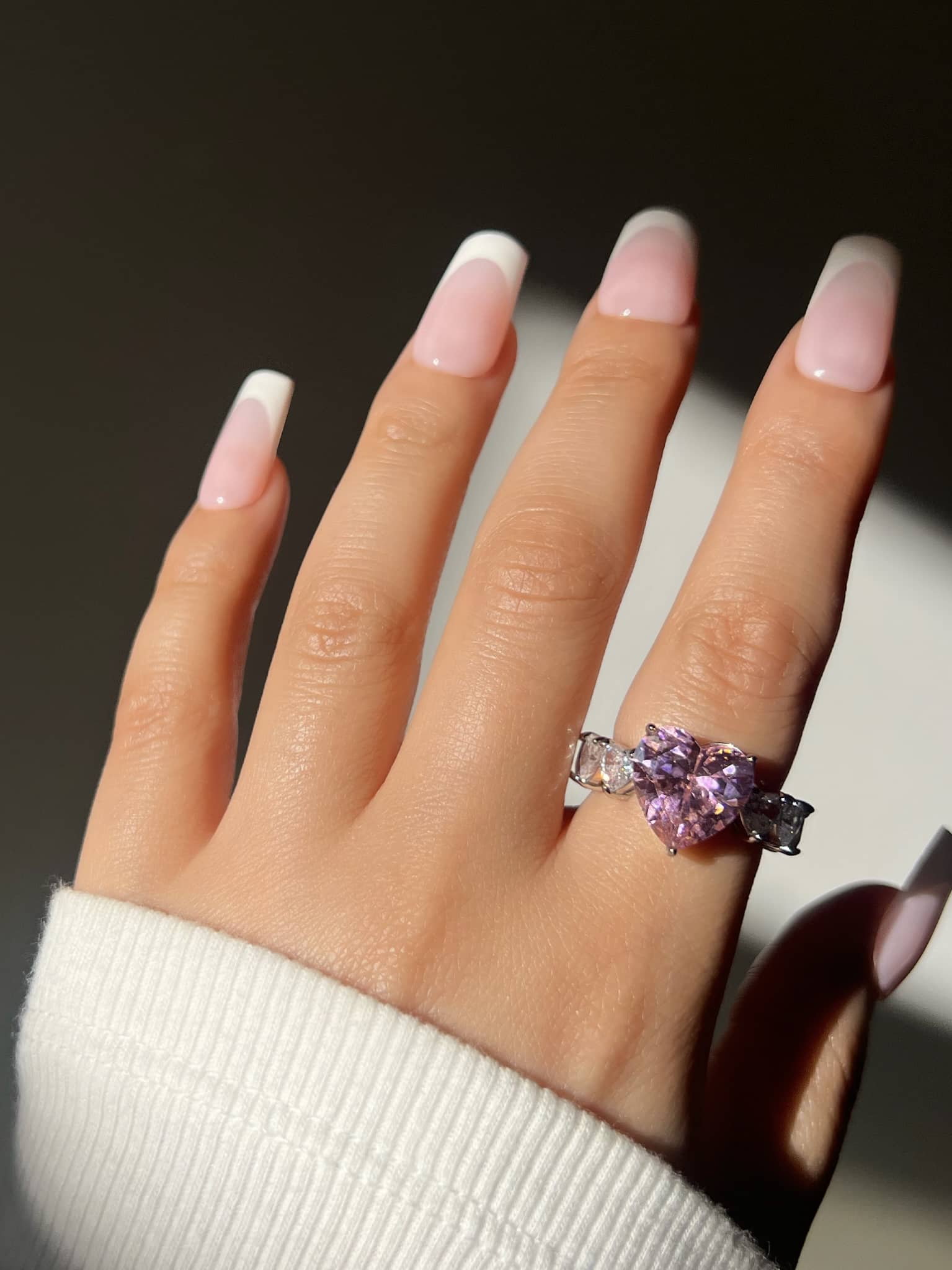 A woman’s hand with French manicured nails showcases a pink and silver ring on the index finger. The ring features a large pink heart-shaped gemstone surrounded by smaller clear stones, highlighting a trendy, elegant, and girly jewellery look.