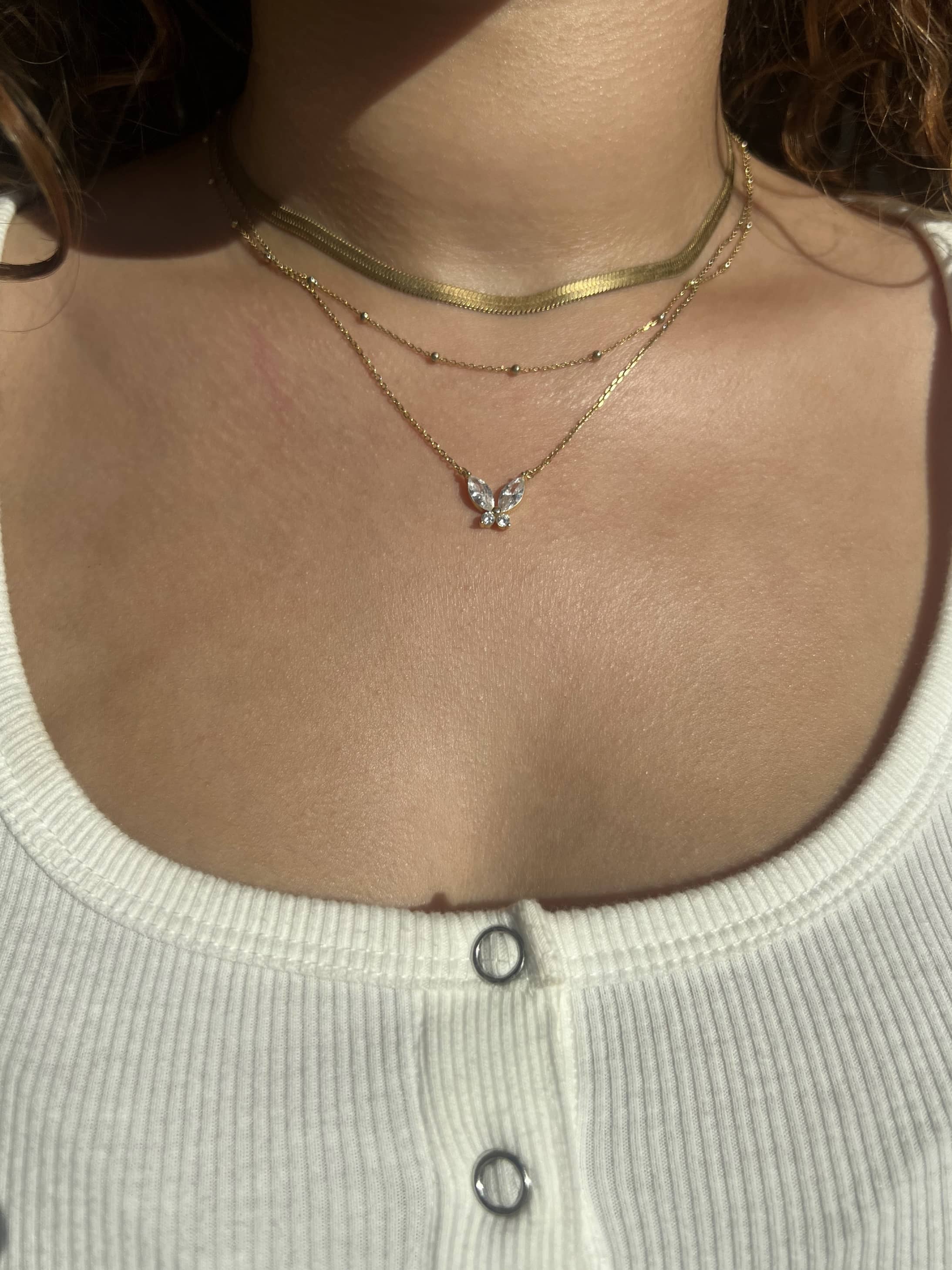 A woman is wearing a white ribbed tank top featuring two silver buttons, complemented by a gold diamond butterfly pendant necklace, a gold snake chain necklace, and a dainty gold ball necklace. The outfit exudes a casual yet stylish vibe.