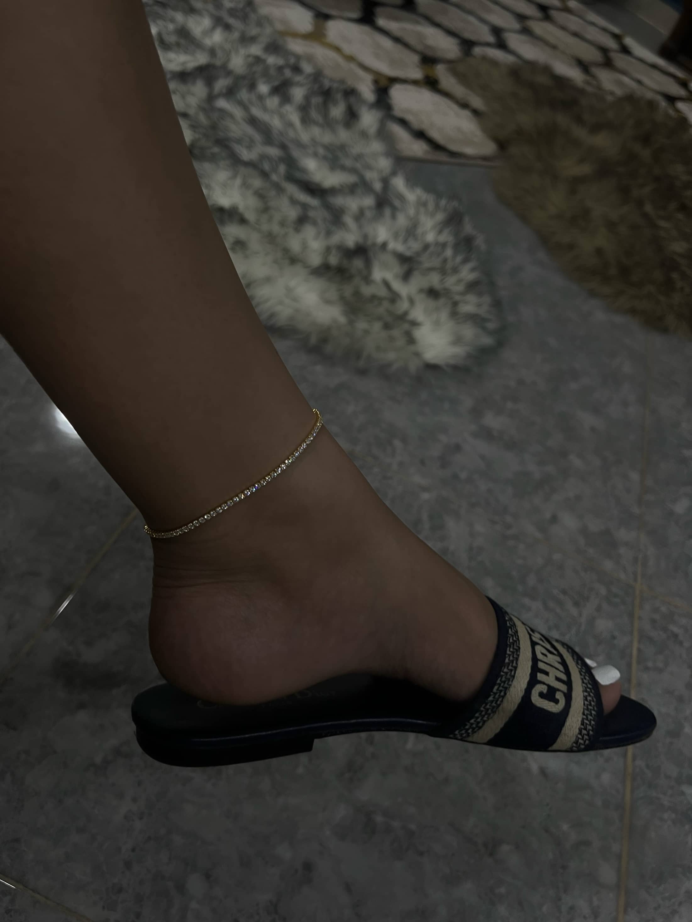A thin gold ankle bracelet adorned with small, clear diamonds, elegantly worn on a woman's foot. The anklet exudes simplicity and sophistication, complementing the navy blue and white "CHRISTIAN DIOR" slide sandal sliders. The background has rugs and tile