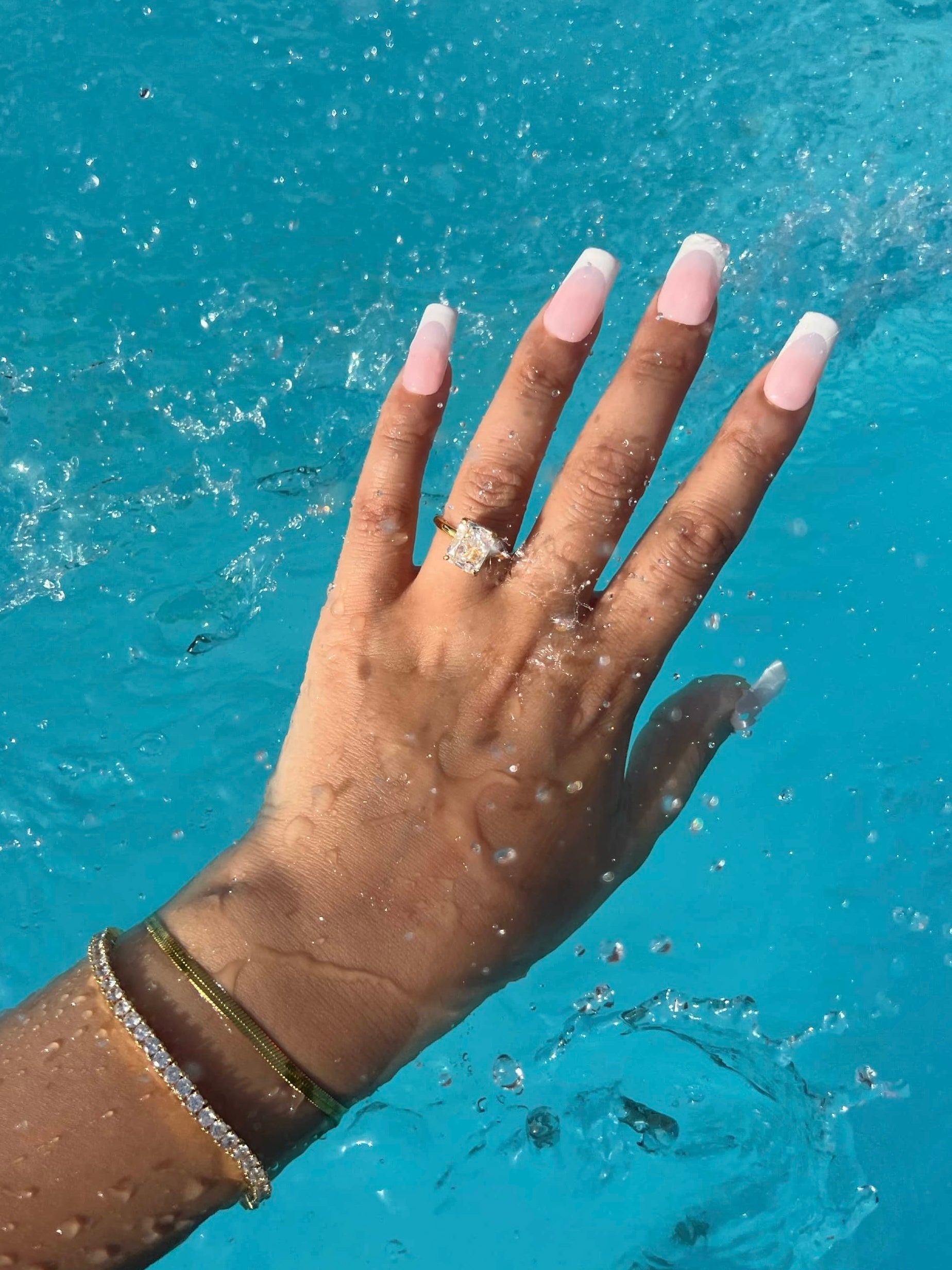 A hand adorned with a large diamond ring and two bracelets is submerged in a swimming pool. The hand features a fresh tip manicure, and the sunlight reflects off the diamond, creating a sparkling effect in the water, showcasing elegance and luxury.