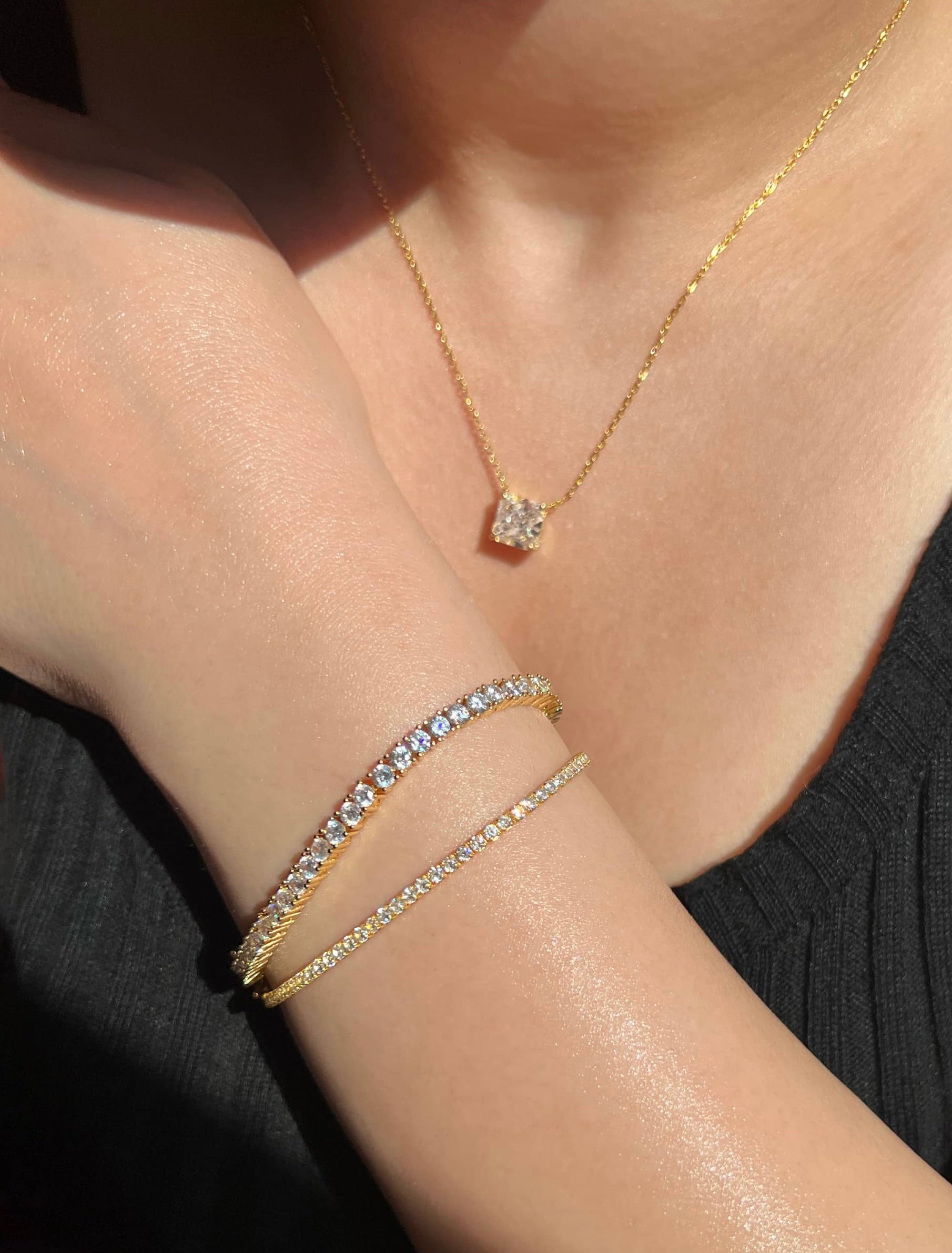 A woman wearing a gold necklace with a square diamond pendant and two gold bracelets. The bracelets feature rows of cubic zirconia diamonds, one tennis bracelet is 2mm and one is 3mm, showcasing a stylish and elegant look perfect for any occasion.