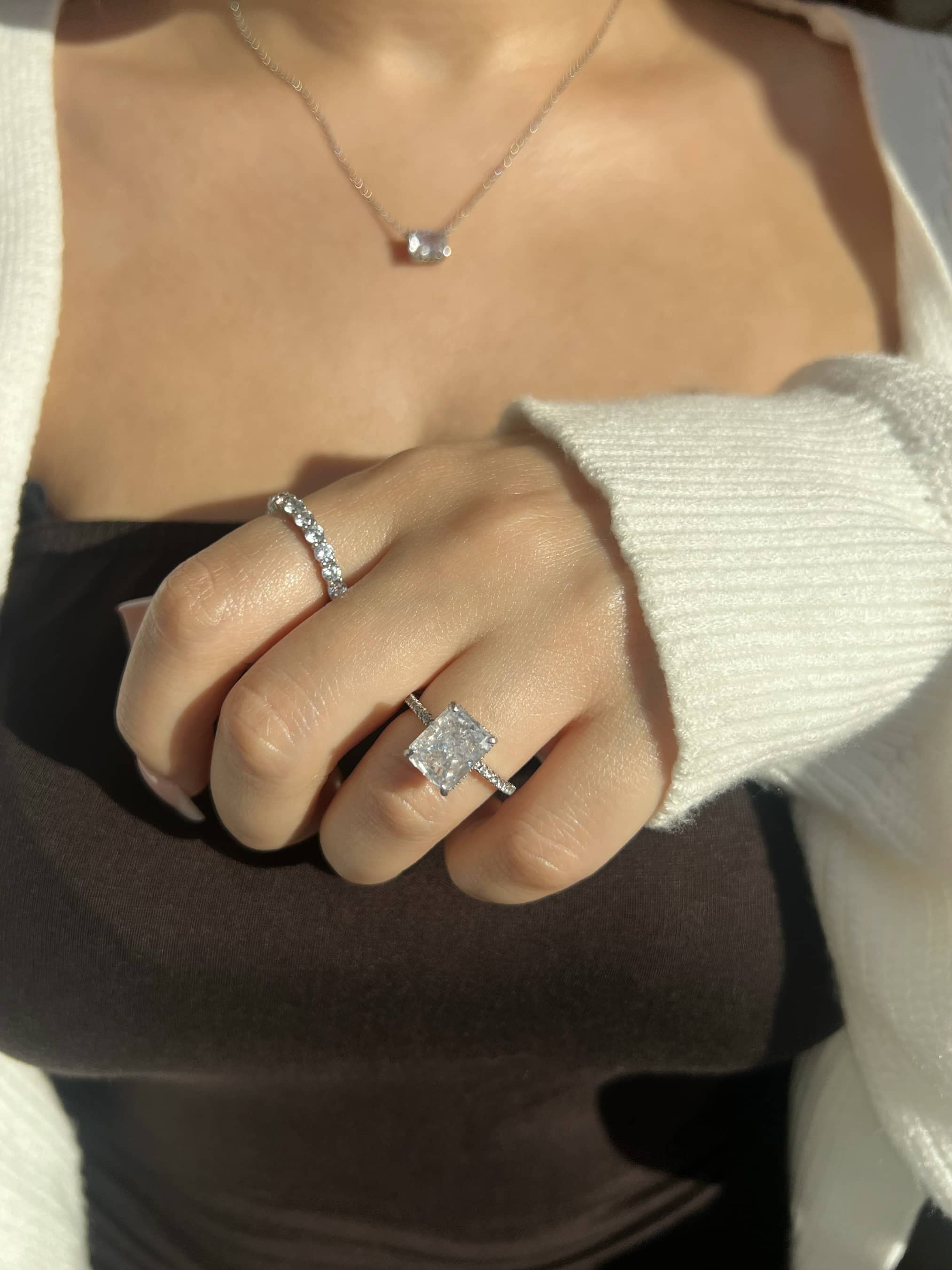 A woman's hand wearing a radiant cut silver ring and a round cut eternity band ring. She is wearing a brown bandeau dress and a white cardigan. She is wearing a radiant cut silver necklace blurred in the background.