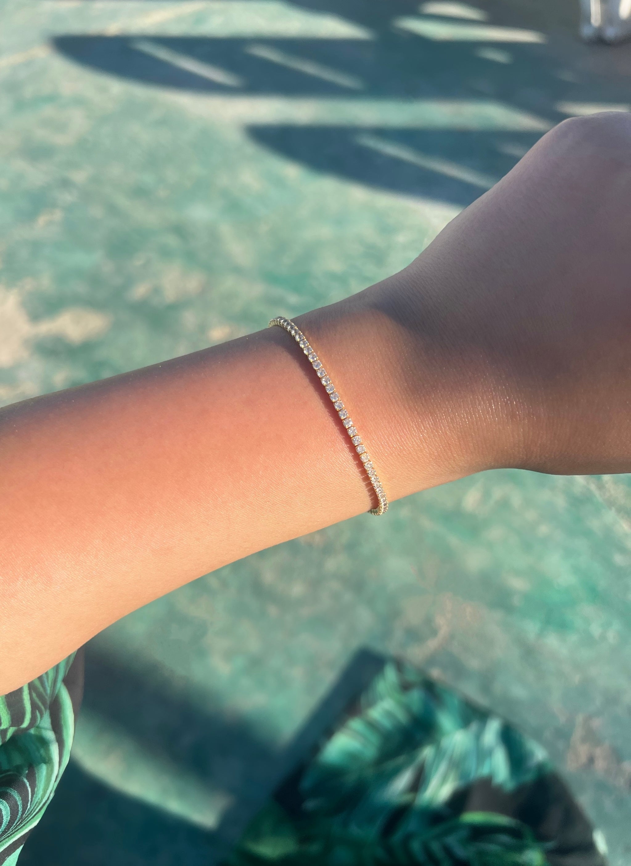 A close-up of a woman’s arm adorned with a thin 2mm gold bracelet featuring small diamonds. The arm rests on a green surface with a floral pattern in shades of green and black, highlighting the elegance of the bracelet against the vibrant background.