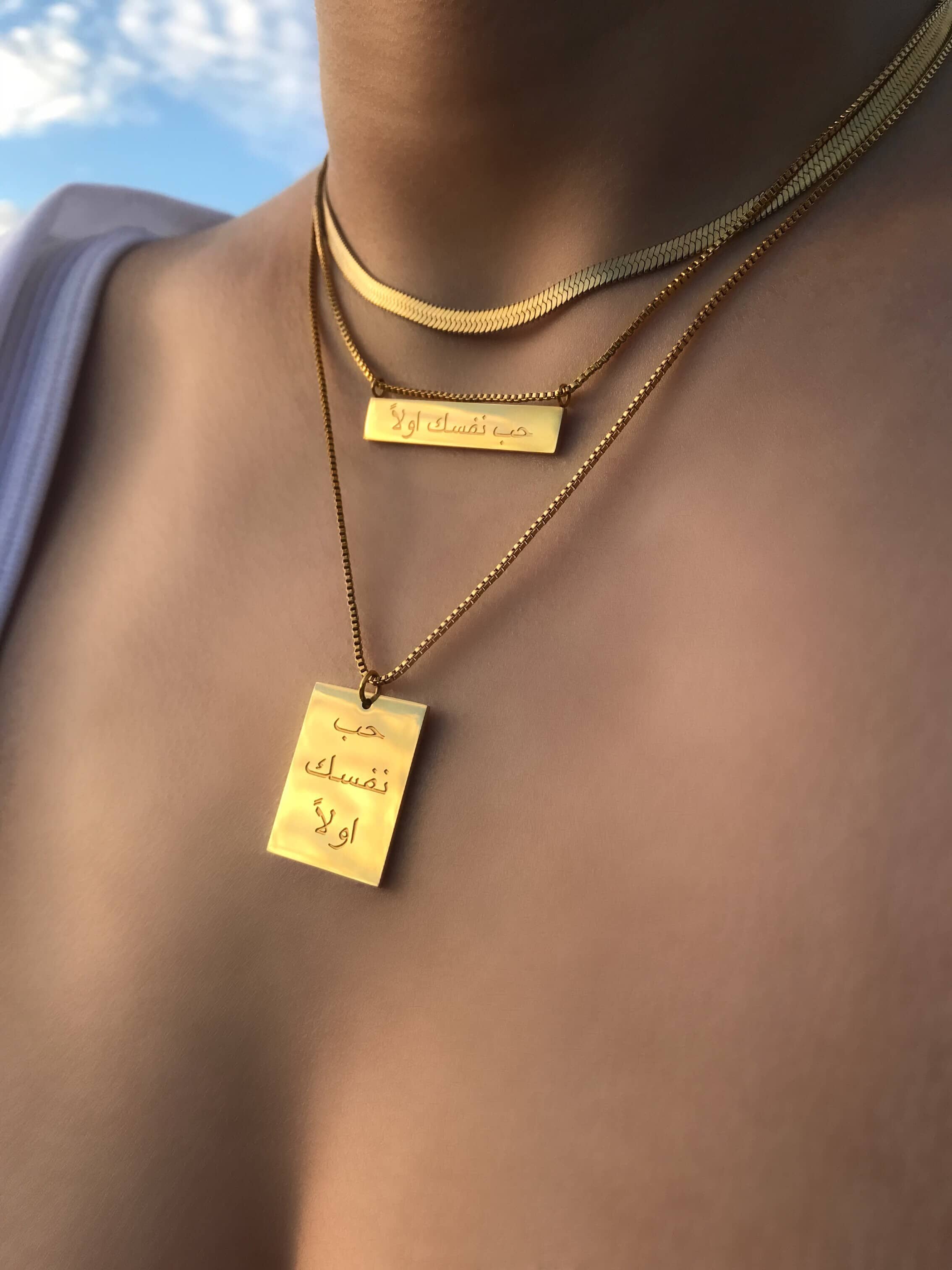 A close-up of a gold herringbone chain necklace with two rectangular pendants. Each pendant is inscribed with "حب نفسك اولا," meaning "Love yourself first," highlighting a beautiful and empowering message. The necklace's shiny finish adds a luxury touch