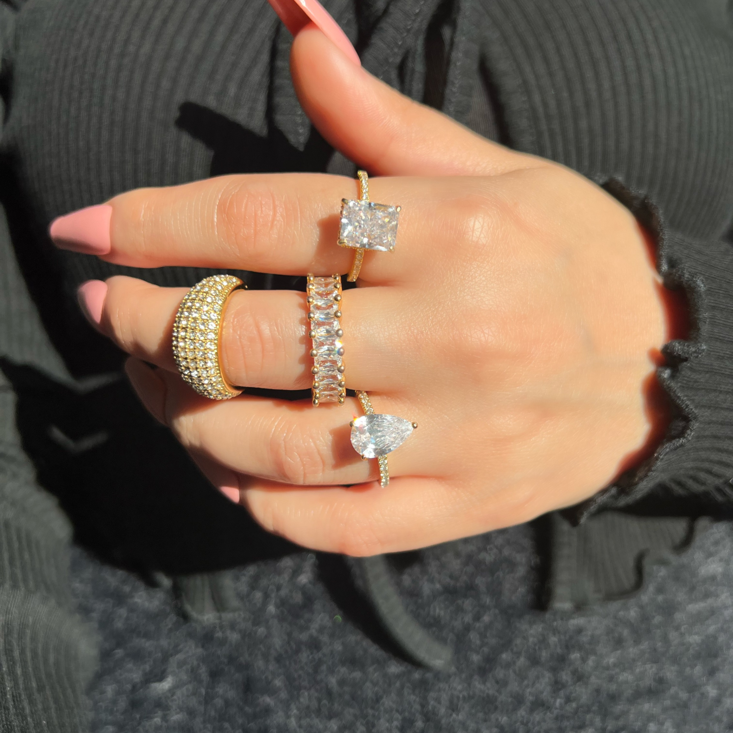 A woman’s hand adorned with four gold rings featuring large clear diamonds. The rings include a large rectangle diamond surrounded by smaller stones, a pear-shaped diamond, and two rings with elegant rectangular diamonds. She has coffin pink nails
