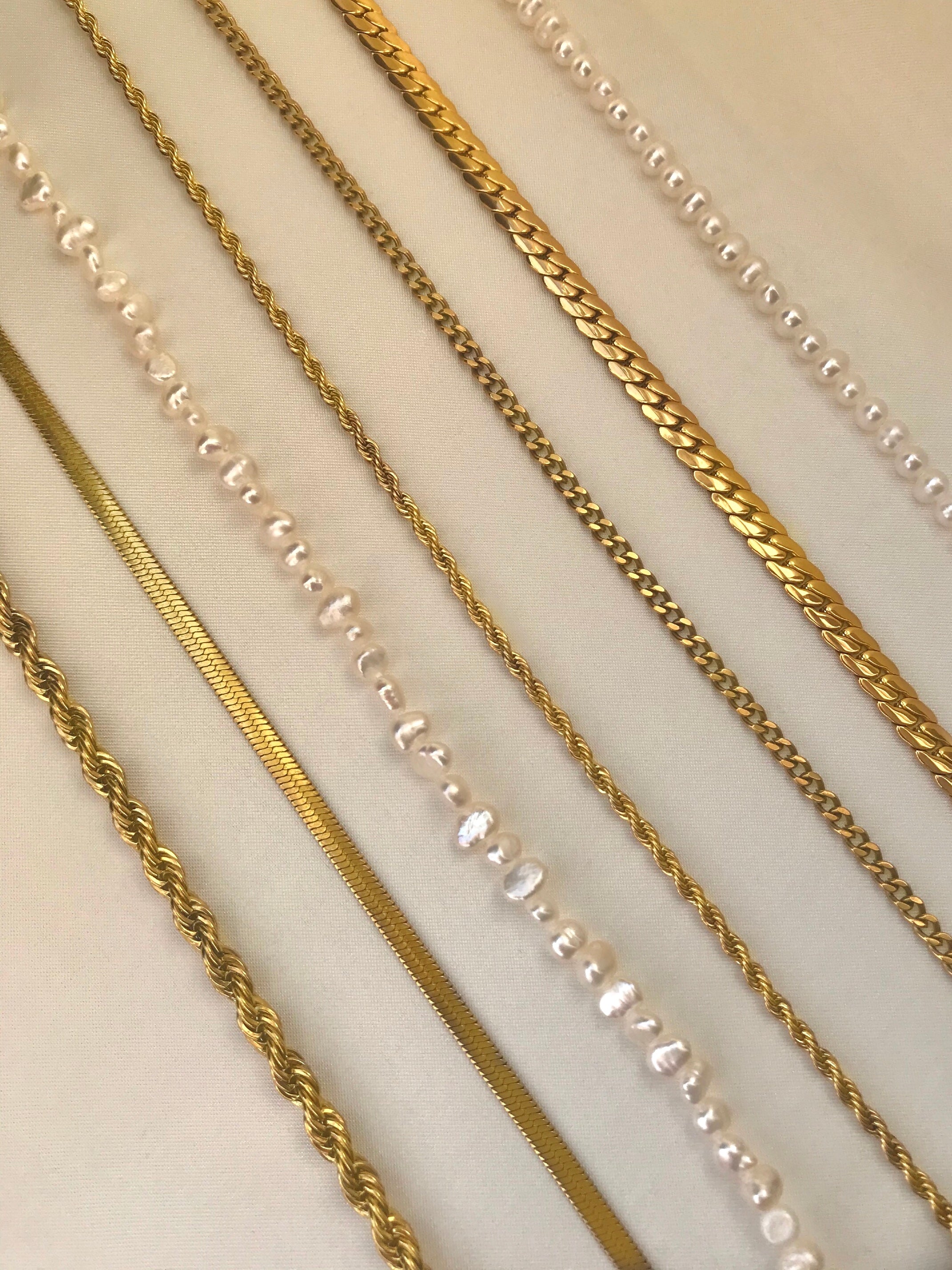 Six gold chains including a rope chain, curb chain, box chain, snake chain, Cuban chain, and delicate chain, and two pearl chains, all displayed elegantly on a beige surface, showcasing their unique textures and styles for versatile jewelry options