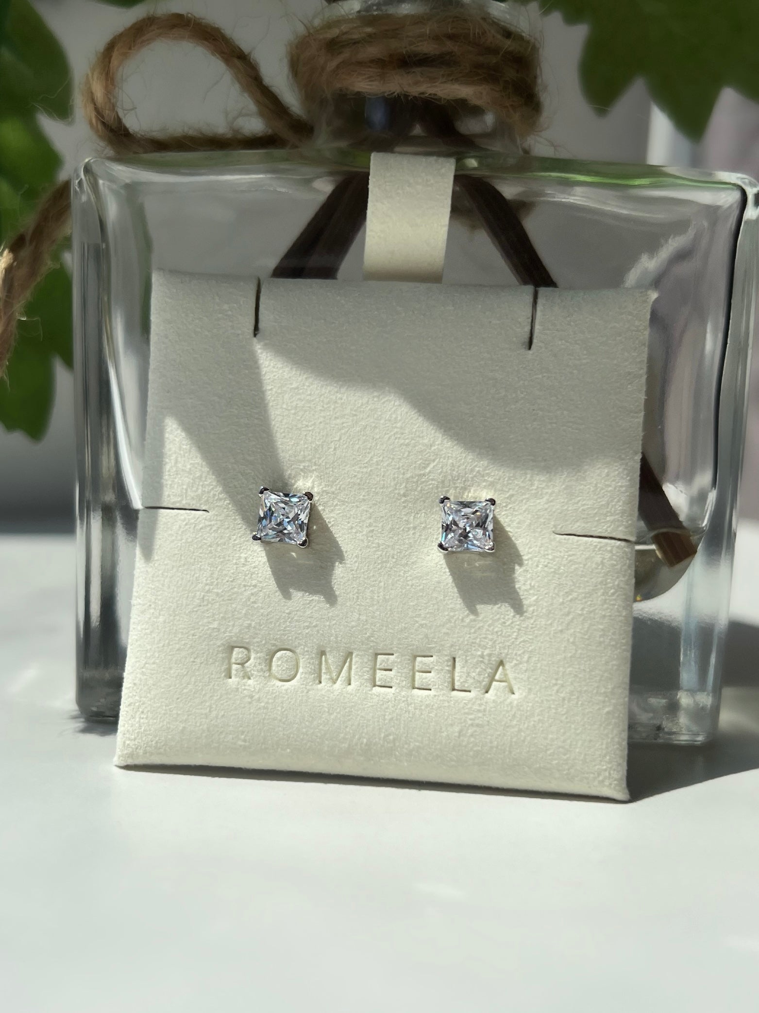 A pair of elegant square-shaped diamond earrings displayed on a white card featuring the brand name 'ROMEELA.' The earrings are crafted in silver, showcasing clear, sparkling stones that catch the light, perfect for adding a touch of glamour to any outfit