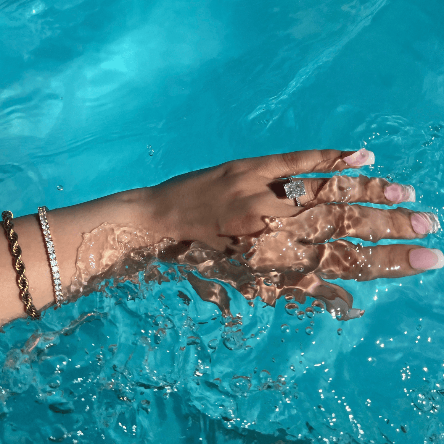 A woman’s hand with long french manicured nails submerged in blue water. The hand features a gold rope chain bracelet, a gold tennis bracelet and a silver radiant cut diamond ring on the ring finger, creating a stunning contrast with the vibrant water