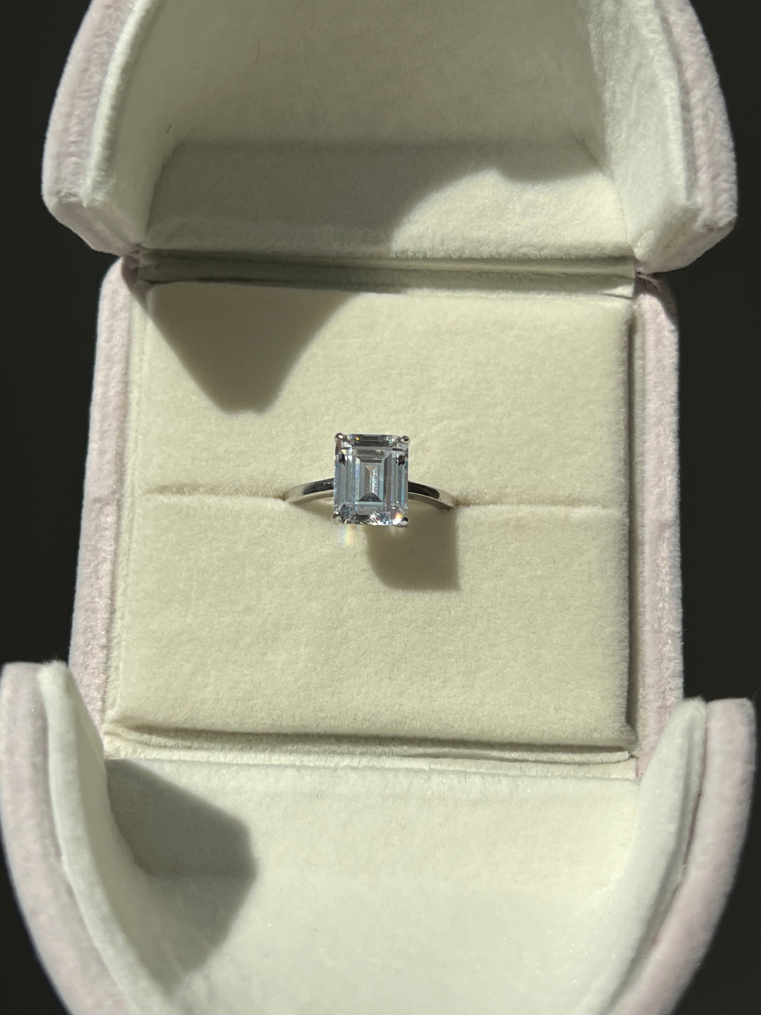 A stunning silver ring featuring a large, clear emerald-cut diamond simulant set in a delicate band. The ring is elegantly displayed inside a light pink velvet ring box, creating a luxurious presentation that highlights its beauty and sparkle