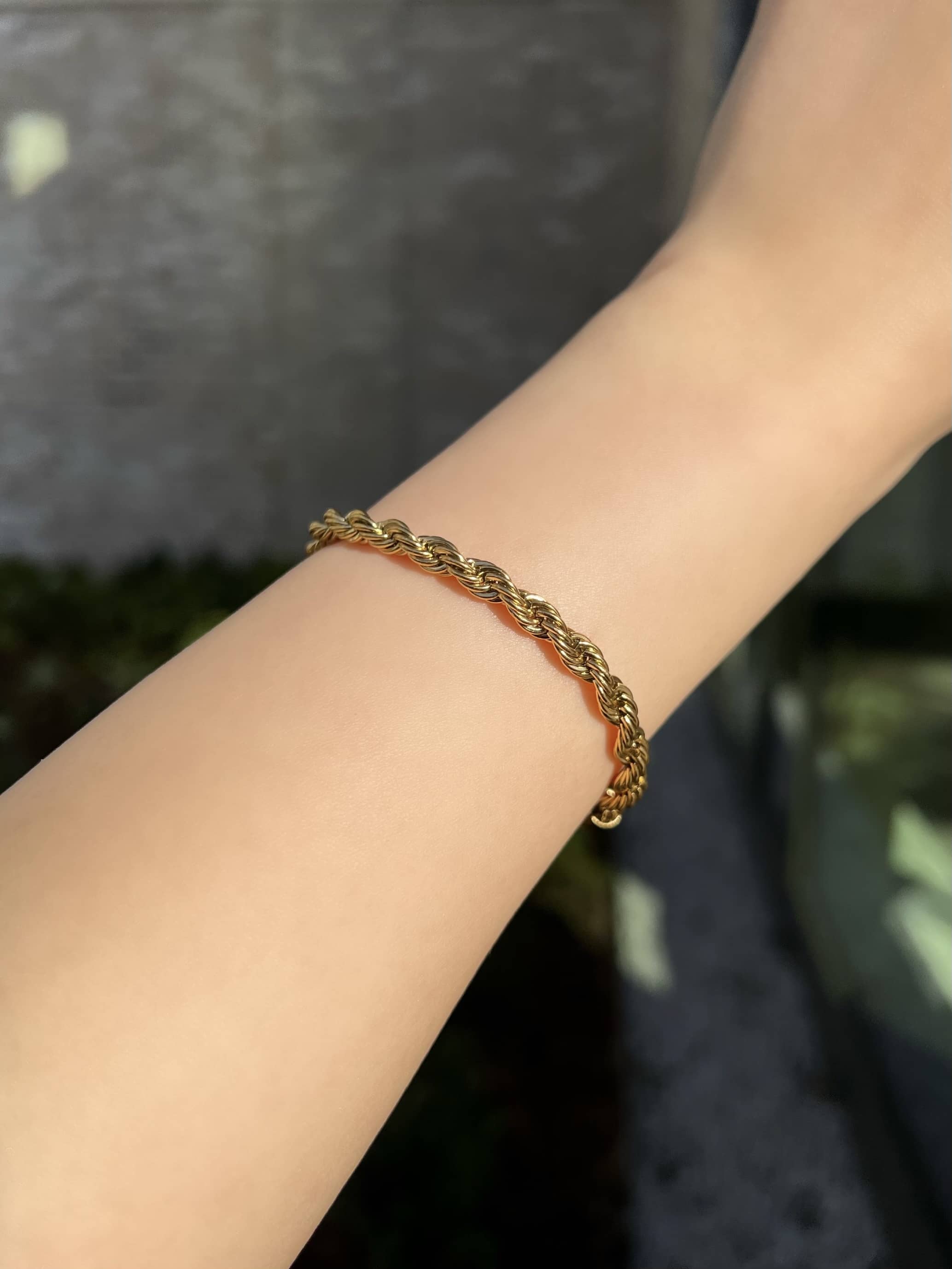 A thick gold rope chain bracelet elegantly adorns a woman’s wrist. The bracelet features a twisted design, showcasing its intricate craftsmanship and adding a touch of sophistication to the wearer's arm. Perfect for both casual and formal occasions.
