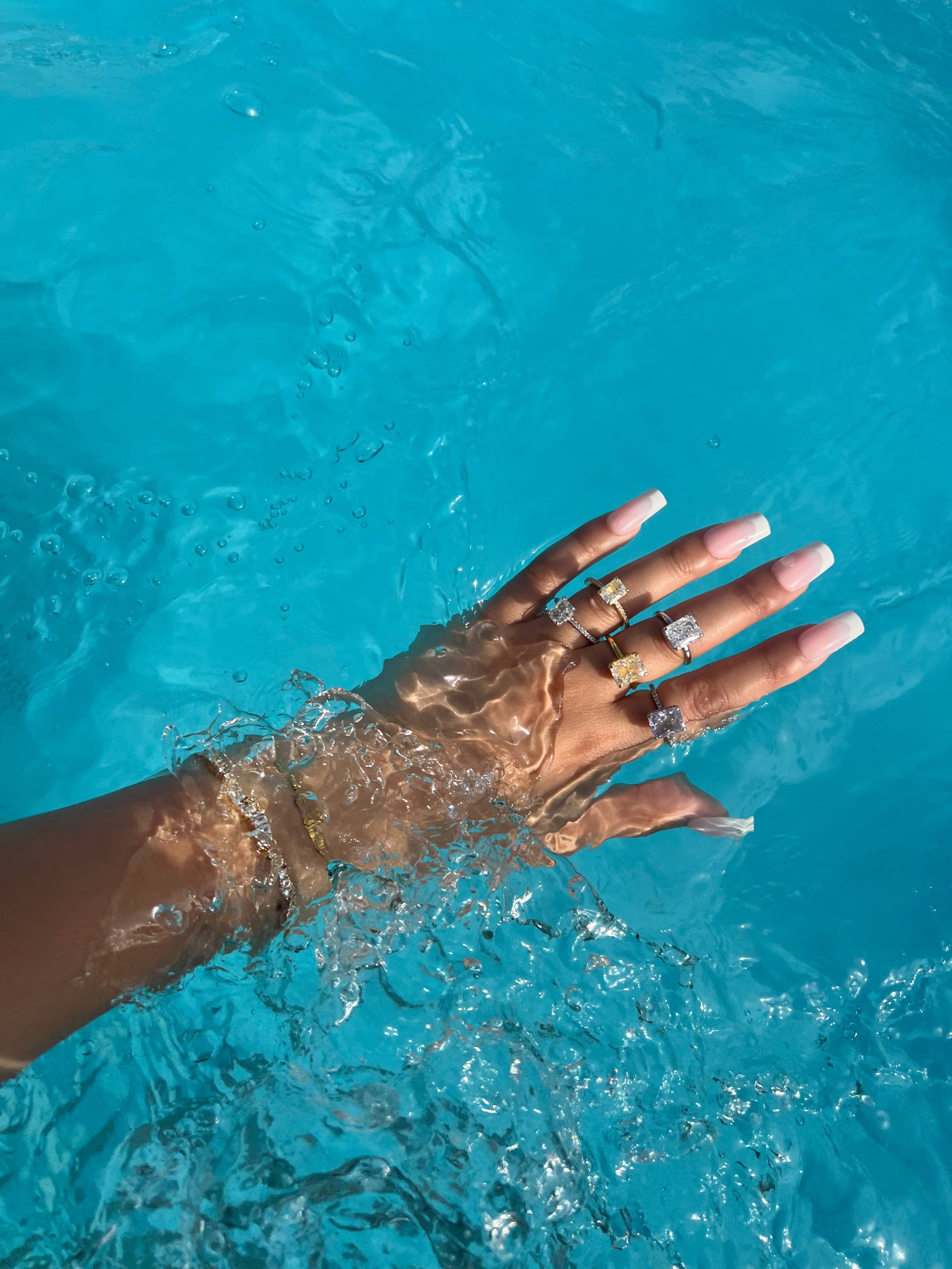 A woman's hand submerged in clear blue pool water, showcasing five diamond rings: one large square yellow diamond and four smaller square clear diamonds set in silver. The hand also features a gold bracelet and long French tip nails
