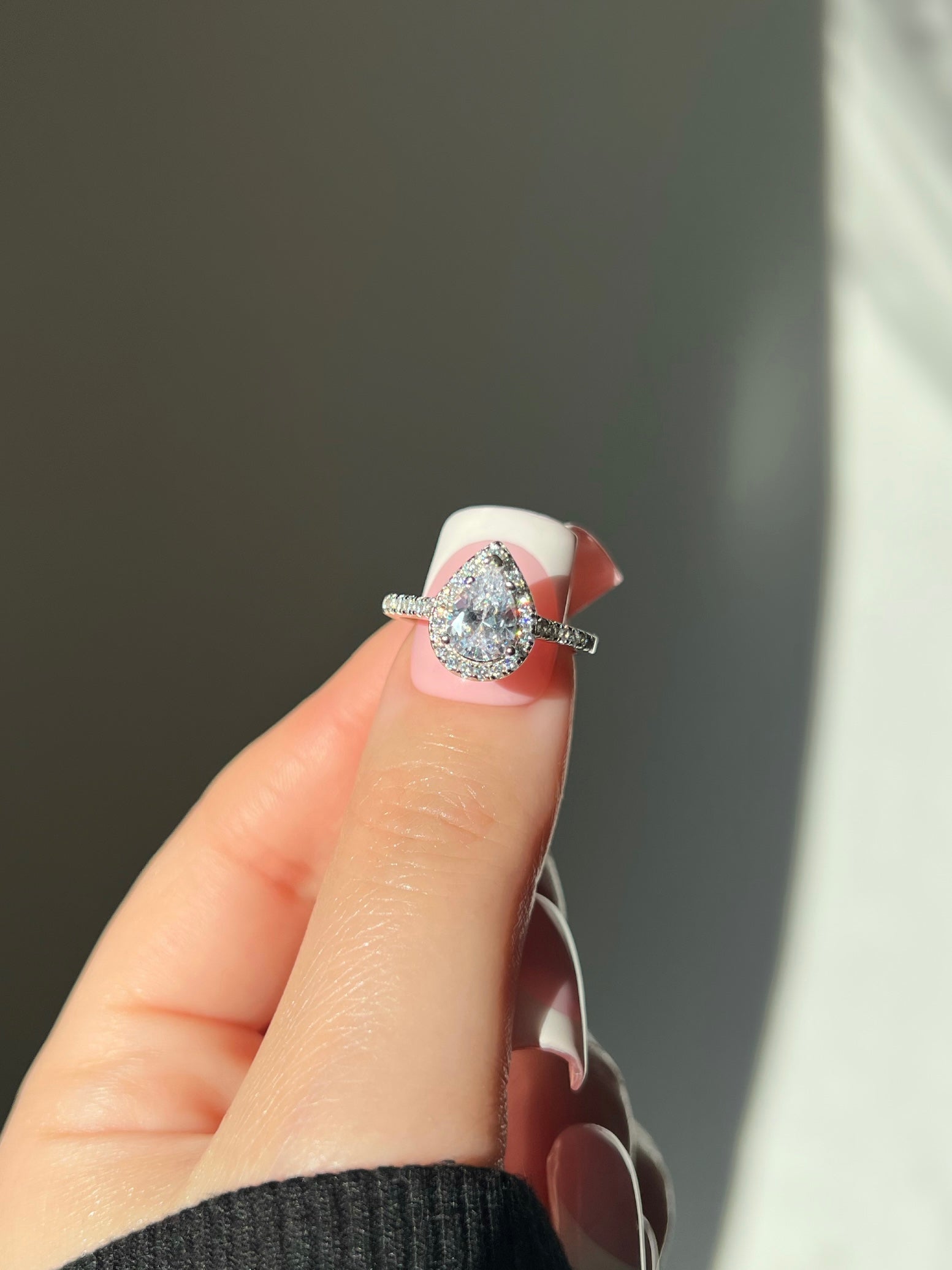 A woman’s hand wearing a stunning pear-shaped silver diamond at the center, surrounded by a halo of smaller diamonds. The ring is set on a diamond-encrusted band, with the hand adorned in a fresh tip manicure, resting on a black sweater. 