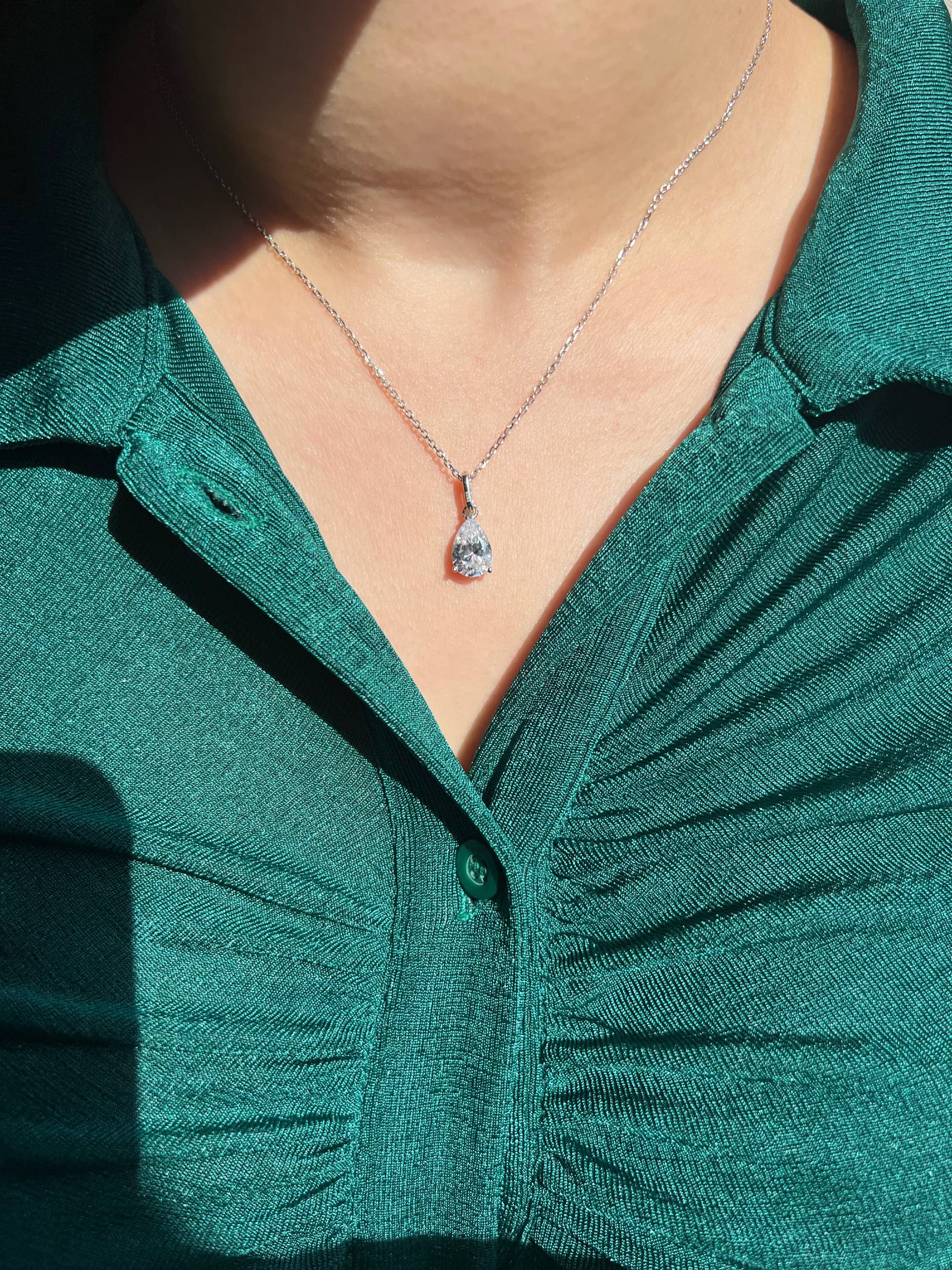 A woman wearing an emerald green top adorned with a stylish silver chain necklace featuring a teardrop-shaped pendant, adding a touch of elegance and luxury to her outfit.