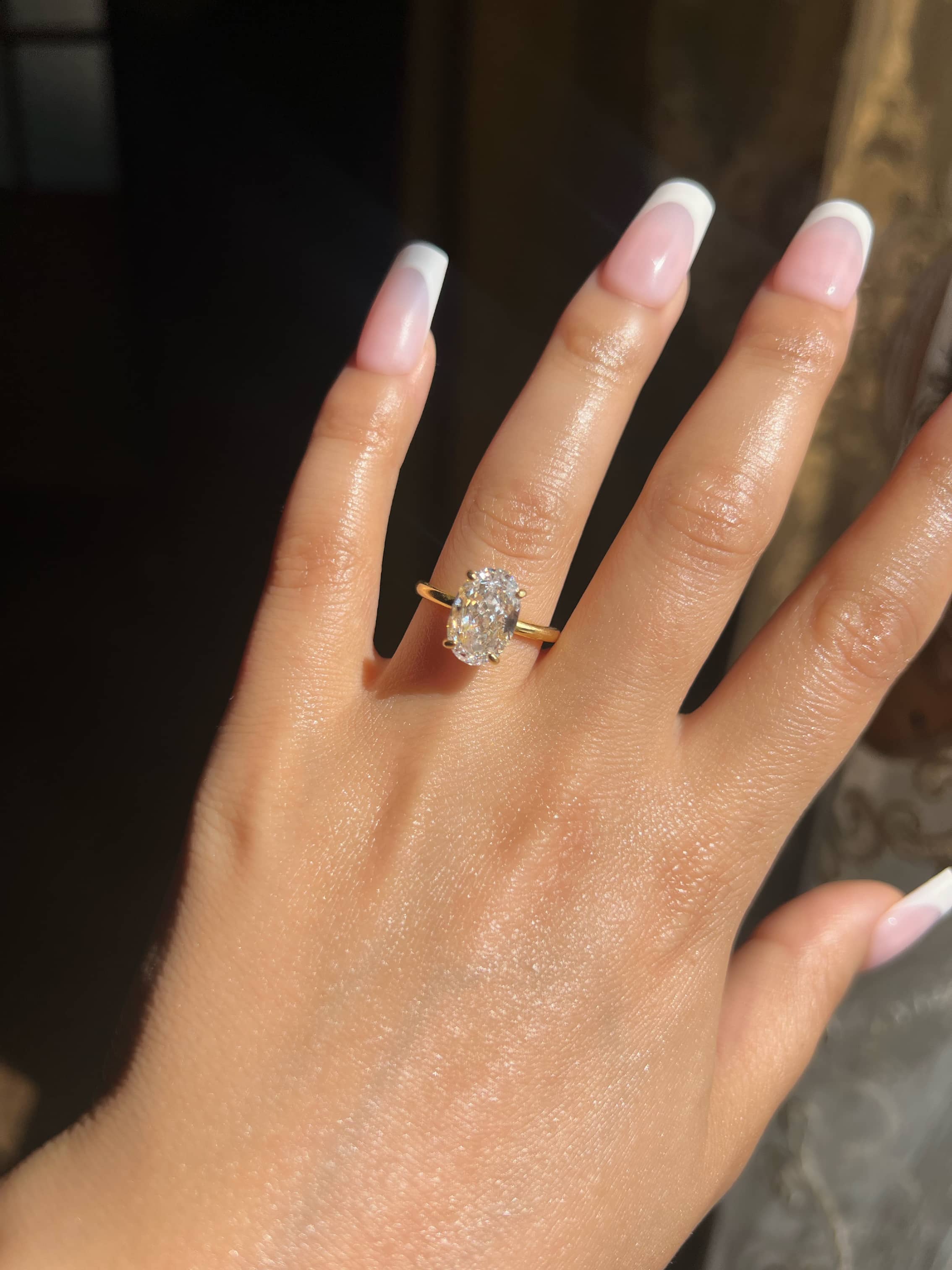 A close-up of a woman's hand showcasing a stunning 3.5 carat oval-shaped diamond engagement ring on her ring finger, complemented by a classic French manicure. The ring features a sparkling crushed ice center stone, radiating elegance and timeless beauty