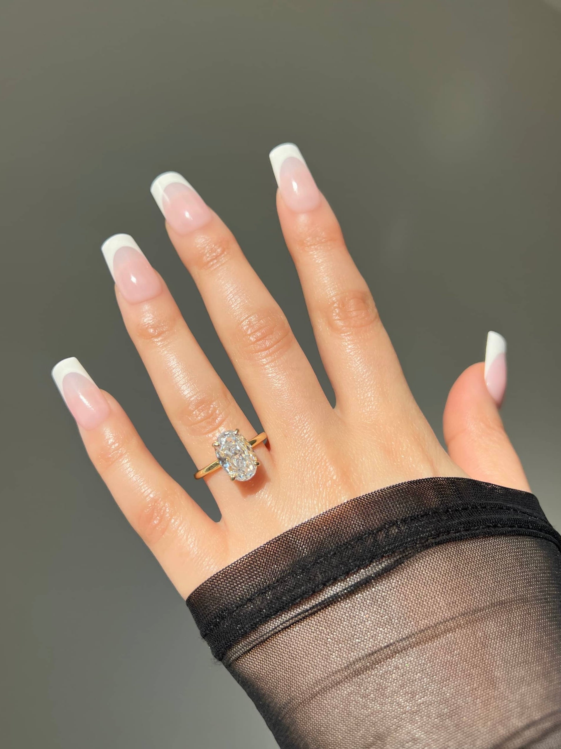 A close-up of a woman’s hand adorned with a beautiful gold oval cut solitaire sterling silver ring.The nails are painted in a chic french manicure. The ring's design highlights the diamond's brilliance, making it a perfect accessory