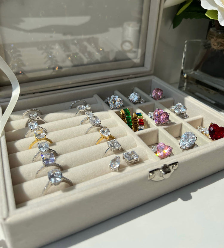 A white velvet jewelry box showcasing an array of stunning rings, including gold diamond rings, a a colourful rainbow ring, pink heart-shaped diamond rings, and diamond simulant engagement rings, all beautifully arranged in a neat grid pattern. 