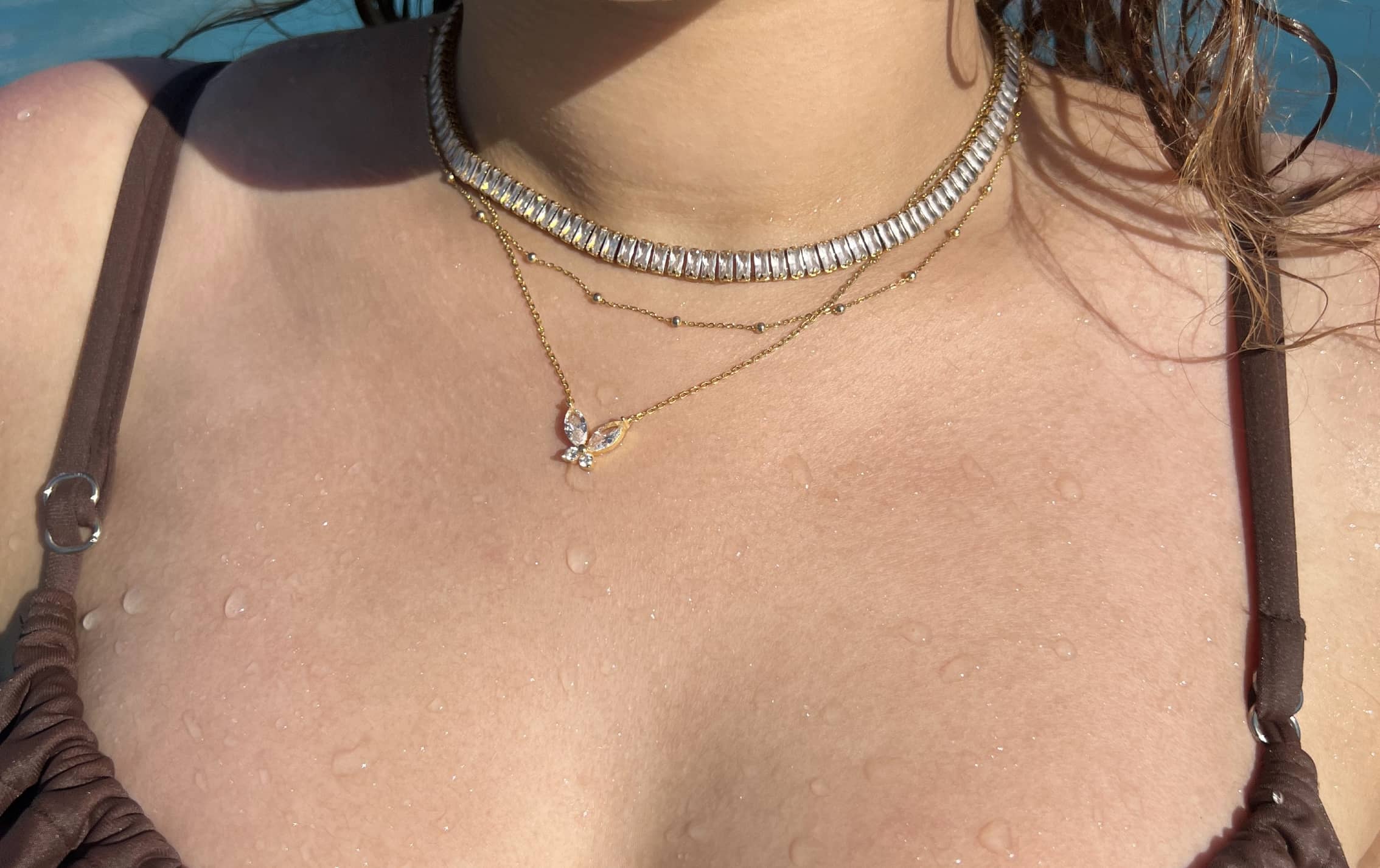 A close-up view of a woman’s chest adorned with three gold necklaces. The top necklace is a baguette tennis necklace, the middle is a simple gold chain with small beads, and the bottom is a delicate butterfly gold necklace, glistening with water droplets