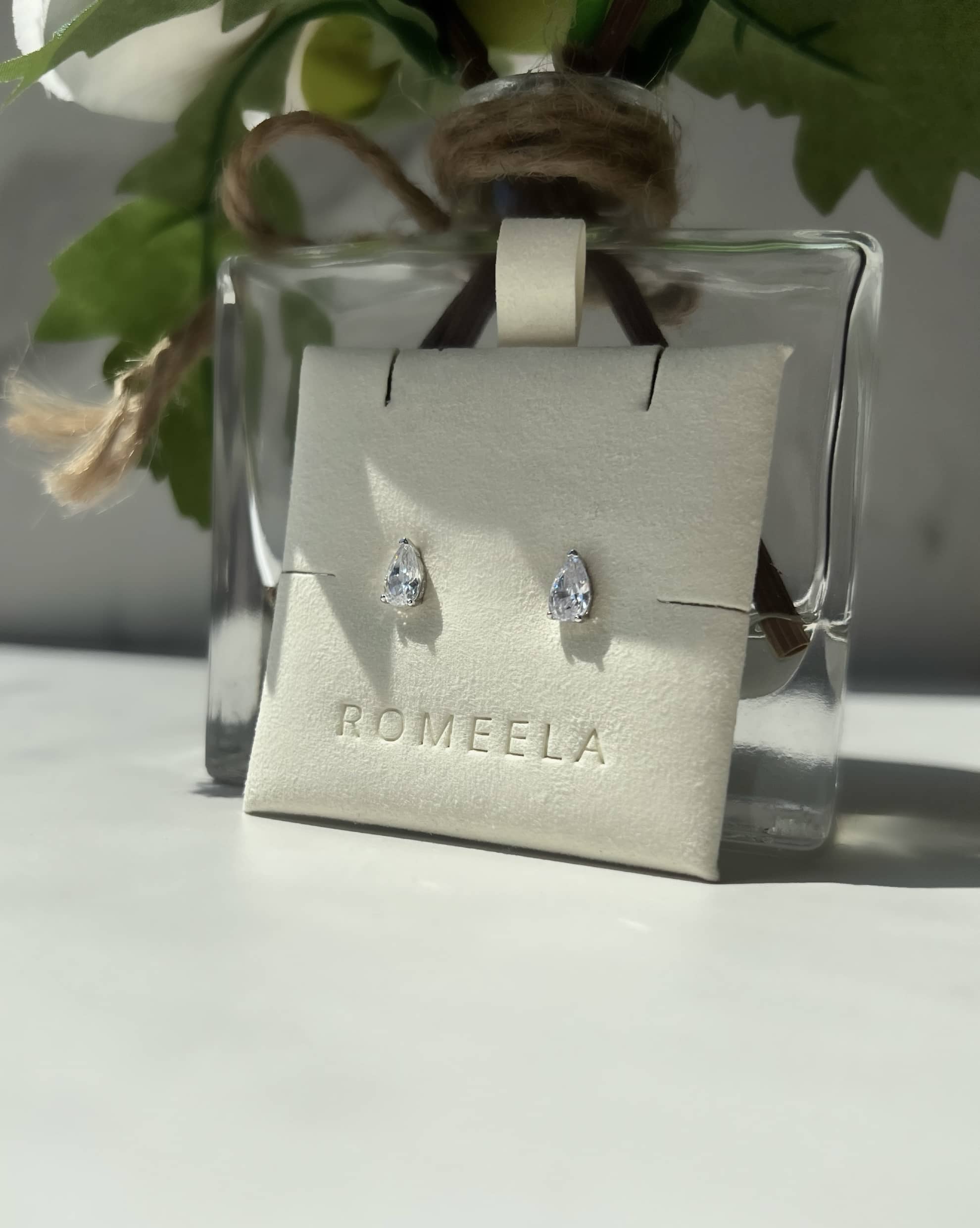 Elegant silver teardrop-shaped diamond earrings displayed on a white card labeled "ROMEELA." The earrings are showcased against a backdrop of a glass vase featuring a white flower and green leaves, highlighting their beauty and sophistication. 