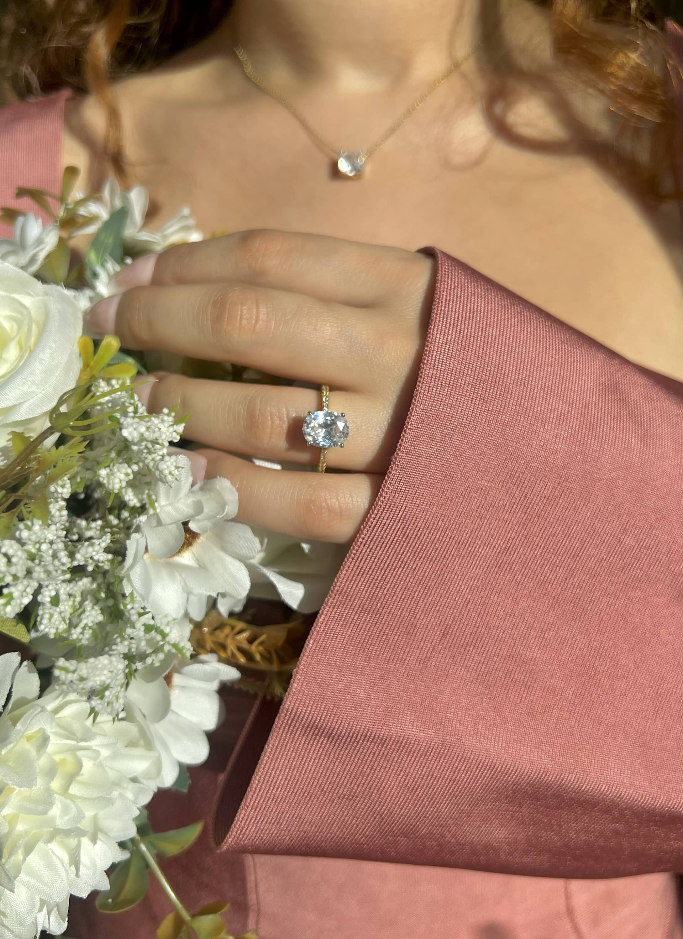 Elegant woman in a pink dress holds a bouquet of white flowers, showcasing a gold 2 carat 8*10mm oval diamond engagement ring, surrounded by smaller diamonds. The ring sparkles beautifully, adding a touch of sophistication to her overall look.