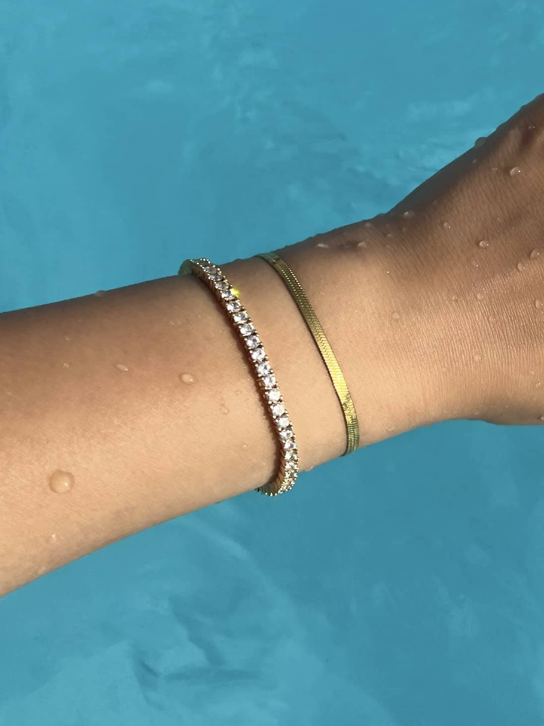 A close-up of a woman's arm adorned with a two gold bracelets; a tennis bracelet and a snake chain bracelet, resting on the surface of a blue pool. Water droplets glisten on the skin, highlighting the elegance of the jewelry against the serene backdrop