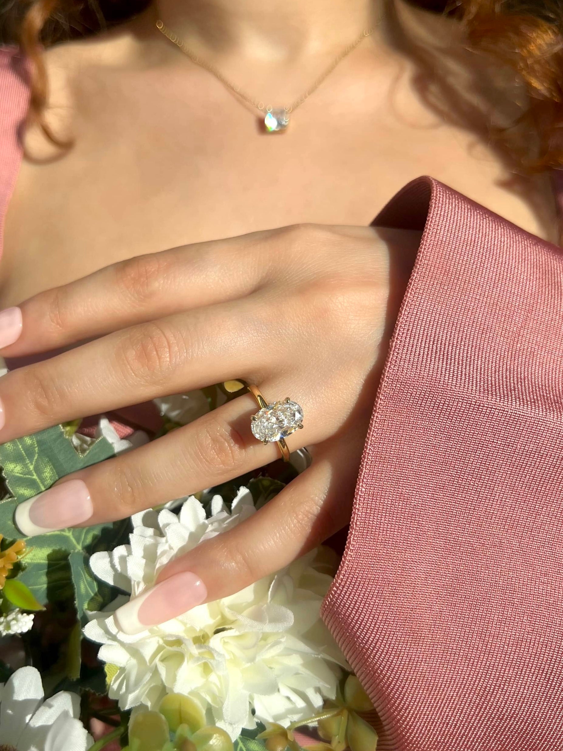 A woman’s hand wearing an oval-shaped 3.5 carat gold diamond ring, resting on a bouquet of white flowers. The hand features long, french tip nails, adorned with a gold necklace with a small, clear stone pendant, all complemented by a pink bandage dress