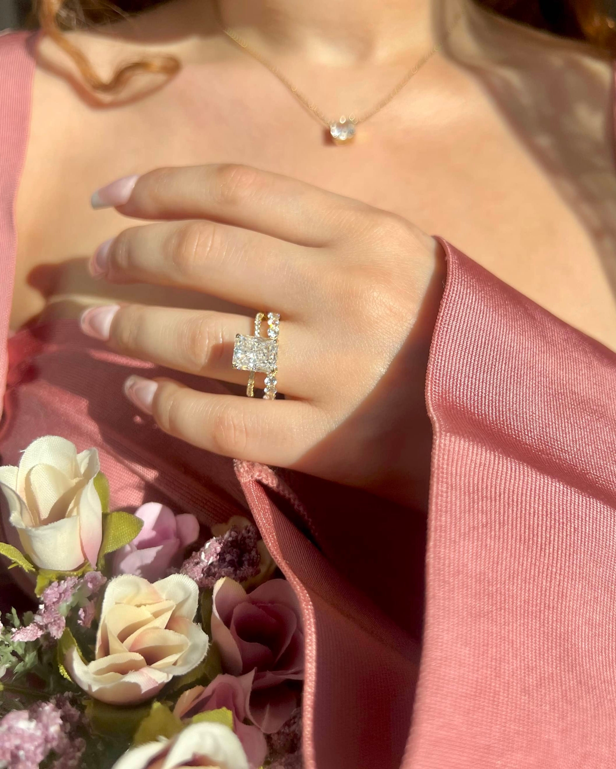 A woman wearing a pink top holds a bouquet of pink roses. She showcases two gold rings on her ring finger: a square-shaped diamond engagement ring and a thinner band adorned with smaller diamonds, adding elegance to her look against the floral backdrop.
